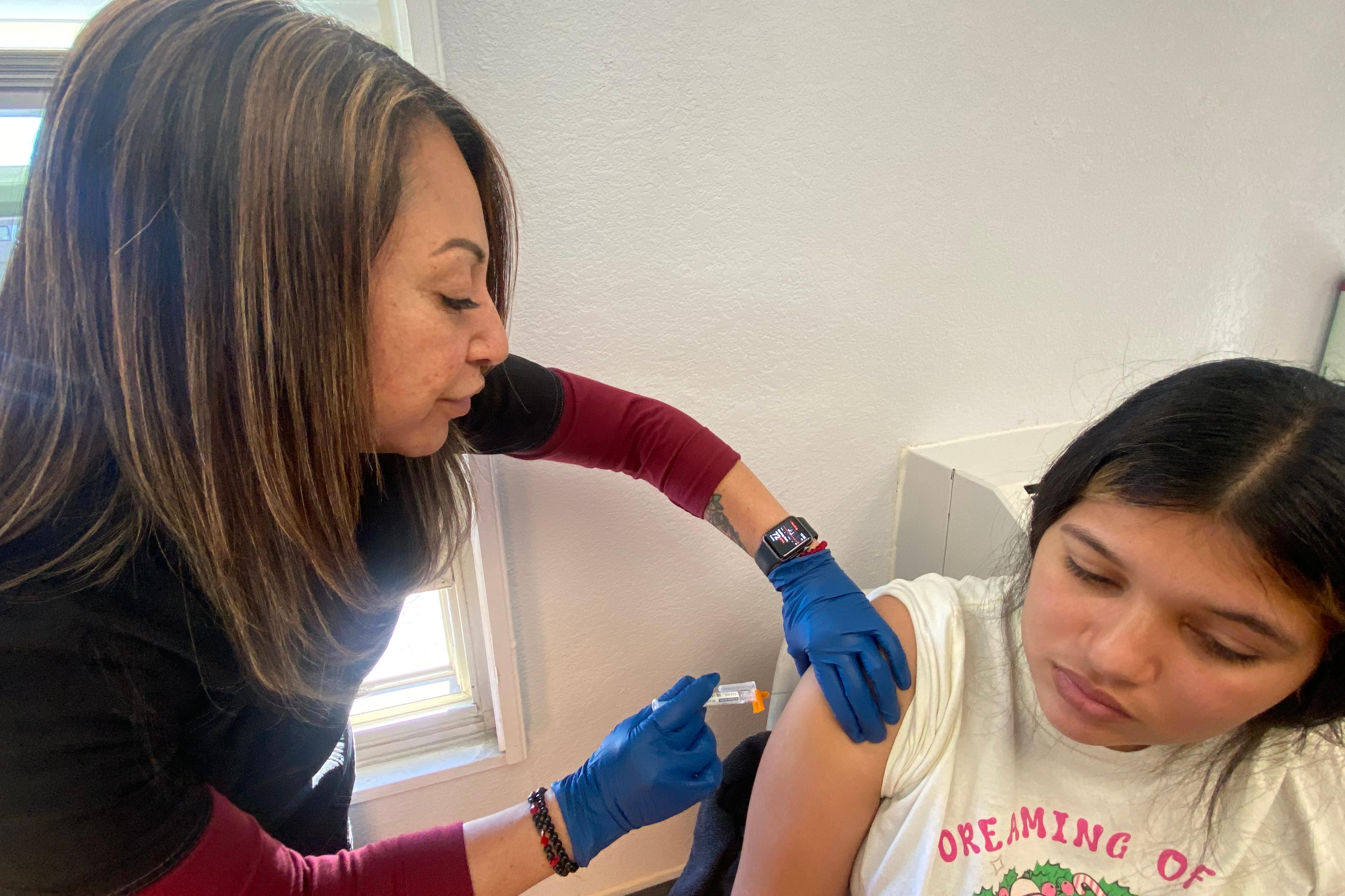 A medical professional wearing black scrubs over a red long-sleeved shirt and blue gloves administers a vaccine to the shoulder of a younger woman.