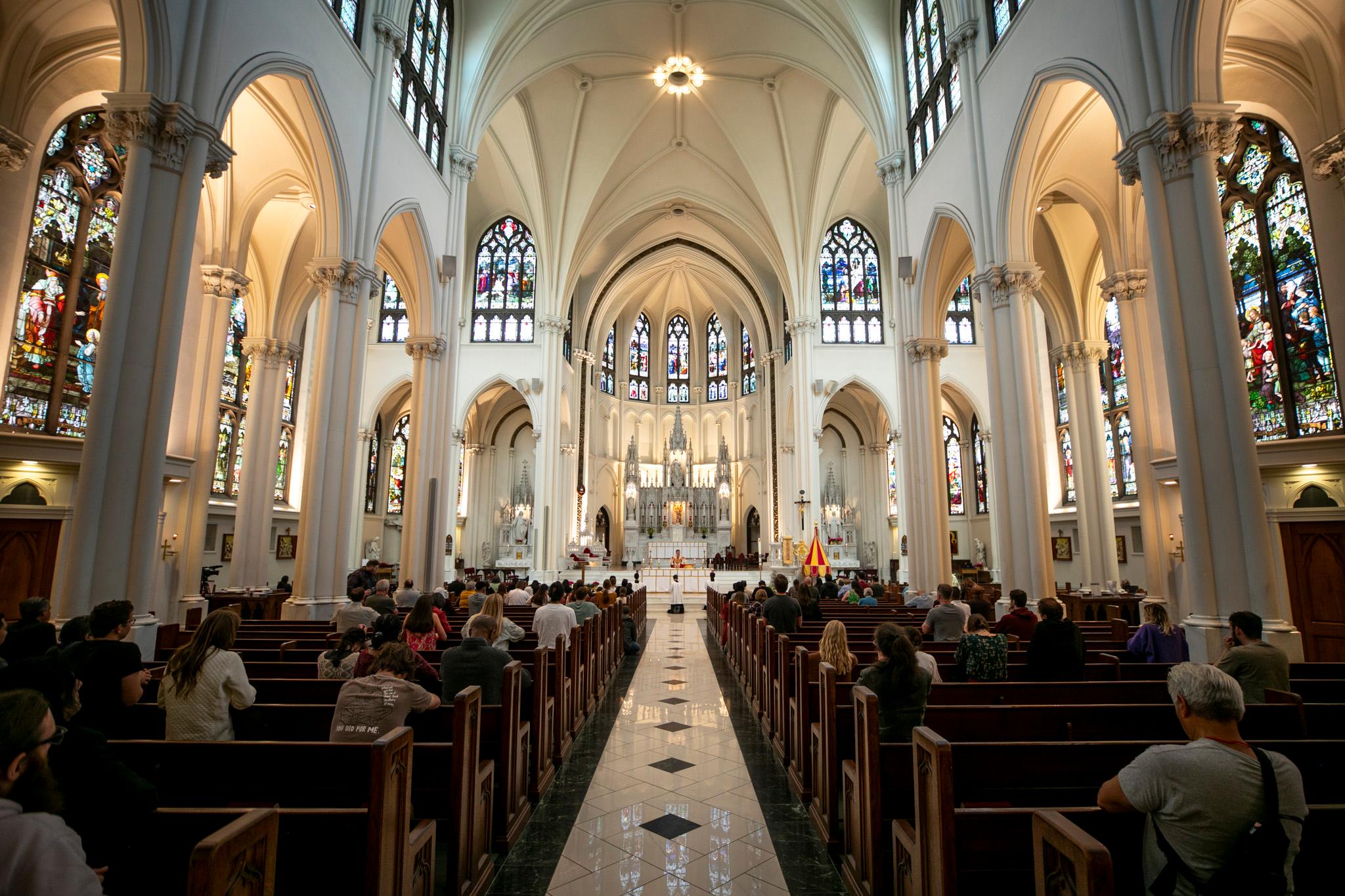 A group of people in pews sit and listen as a priest in the far distance speaks at a lecturn.