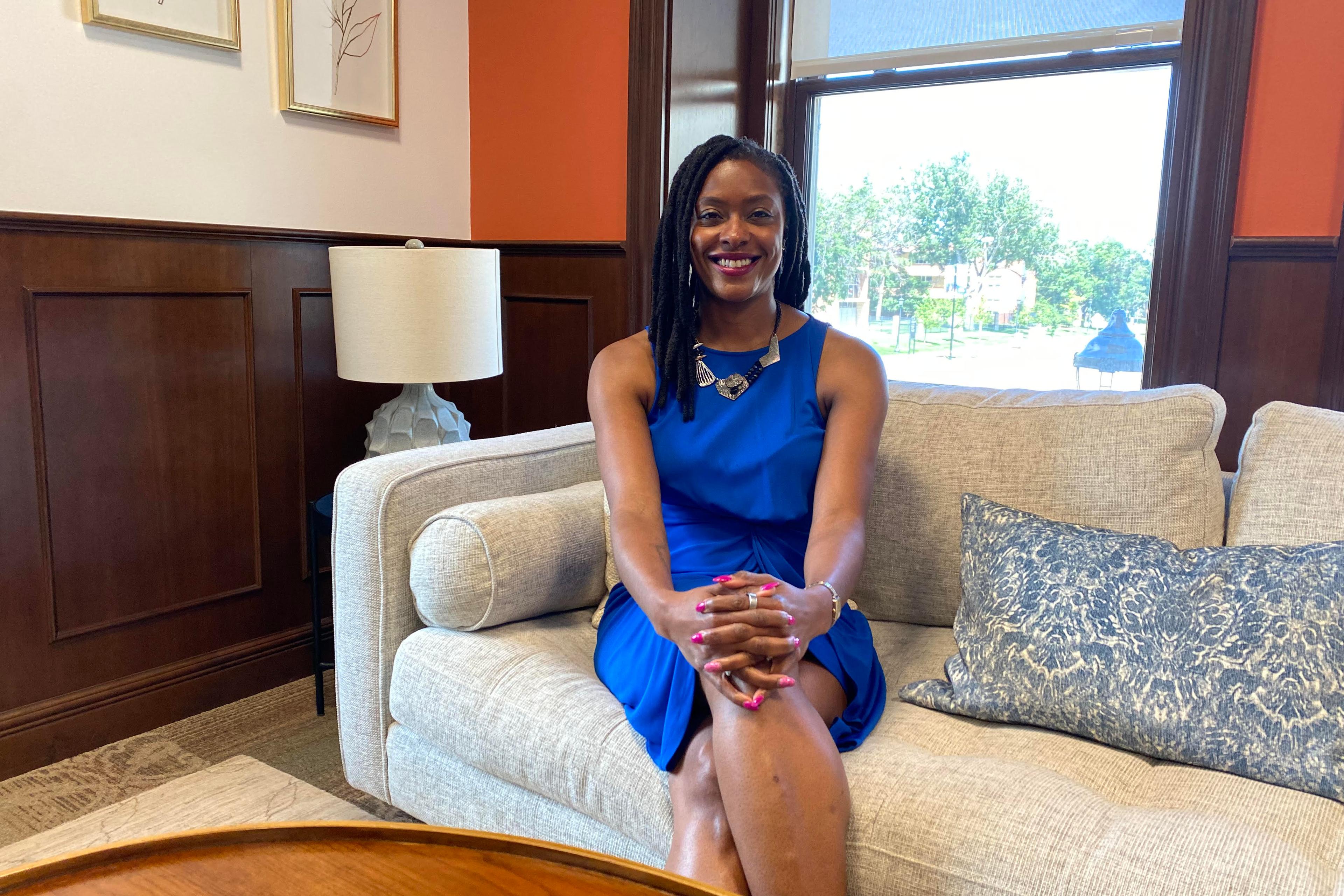 A smiling Manya Whitaker sits on a couch wearing a blue dress.