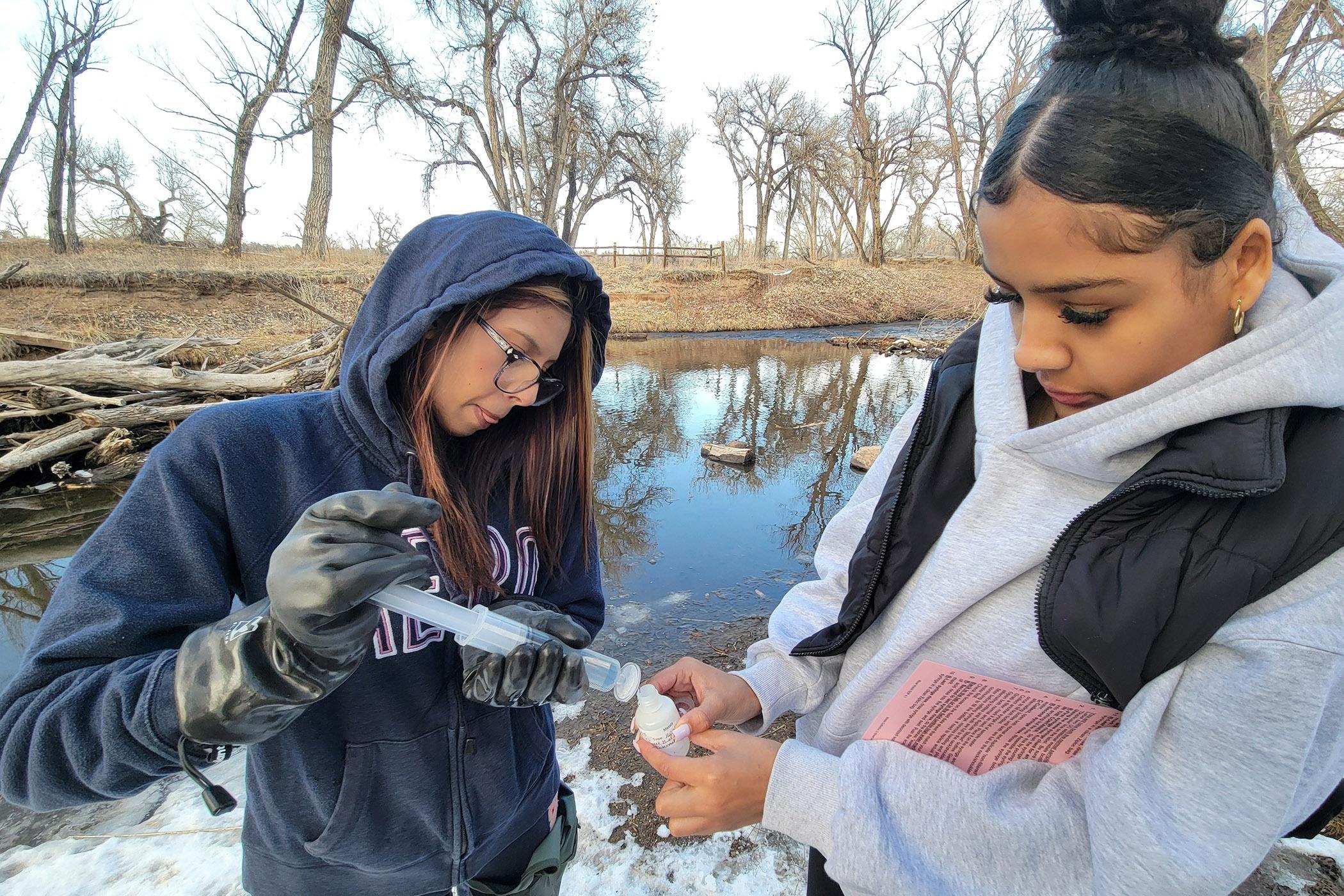 Two girls stand at a snowy bank of a creek. One girl is using a large liquid syringe to put a sample of water into a test container the other girl is holding.