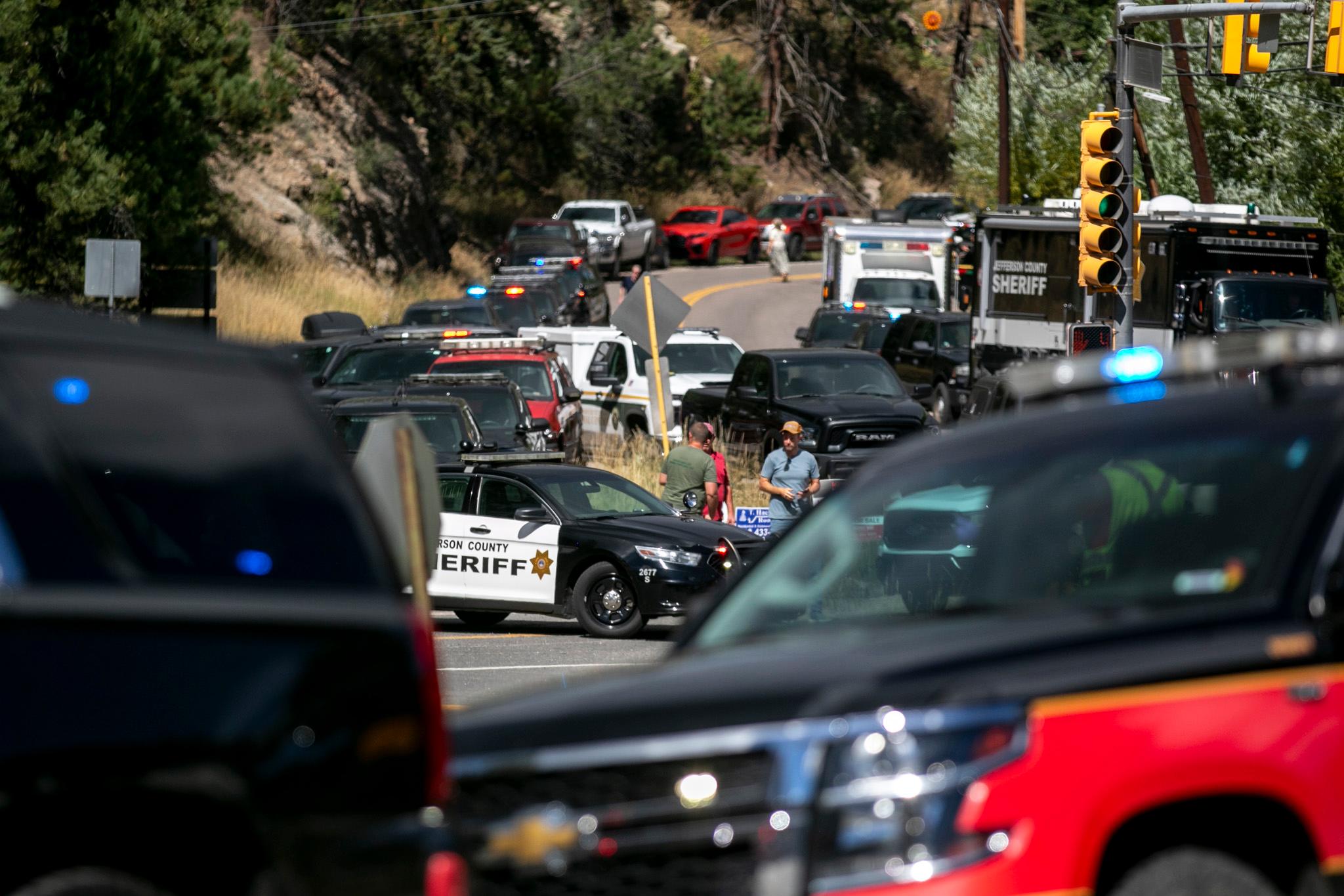 Several police cars and other emergency vehicles block a road lined with trees. Officers and people stand near a county sheriff car.
