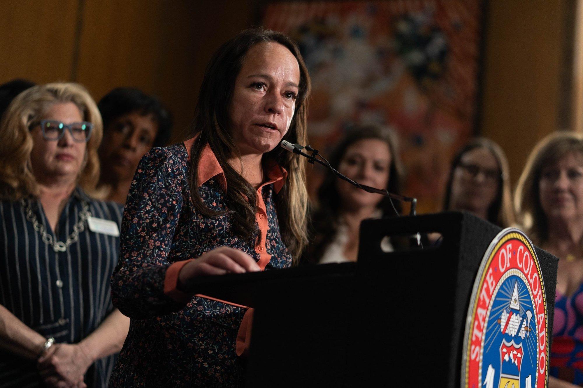 A woman with many other women standing behind her at a podium speaking into a mic