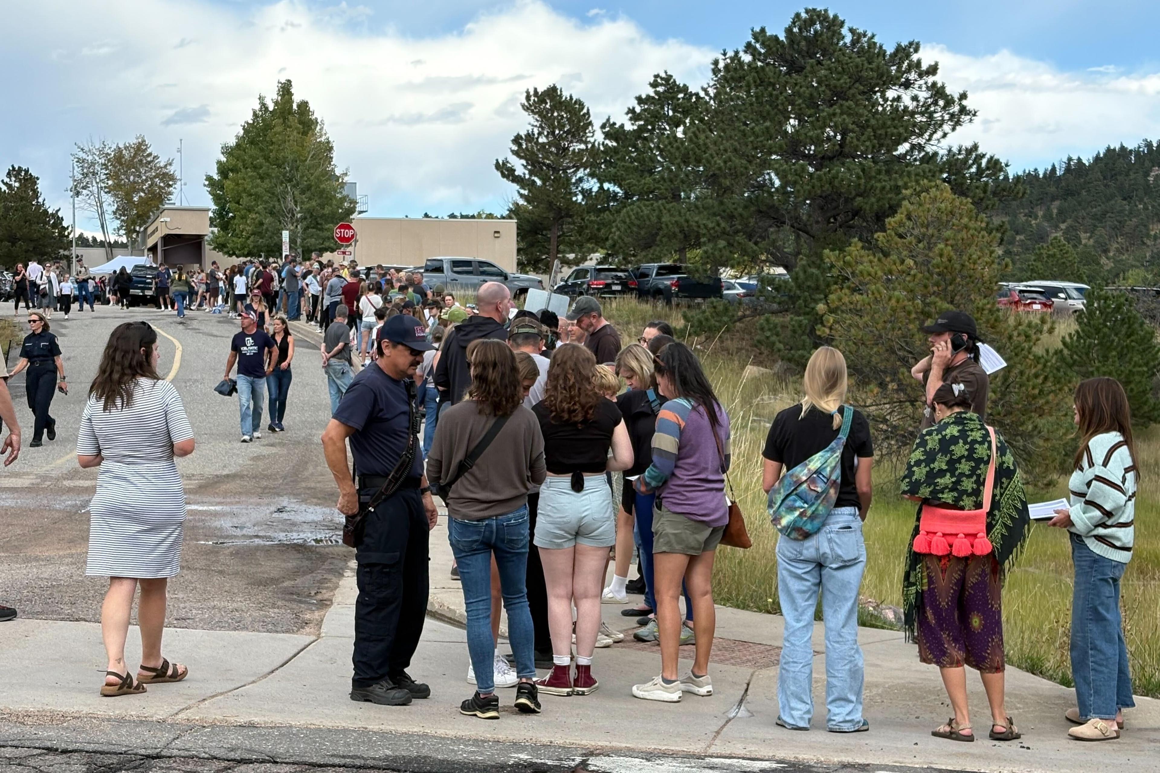 A long line of people stands and waits outside near trees and parked cars on a cloudy day. Some people talk to uniformed officers, while others look ahead or use their phones.