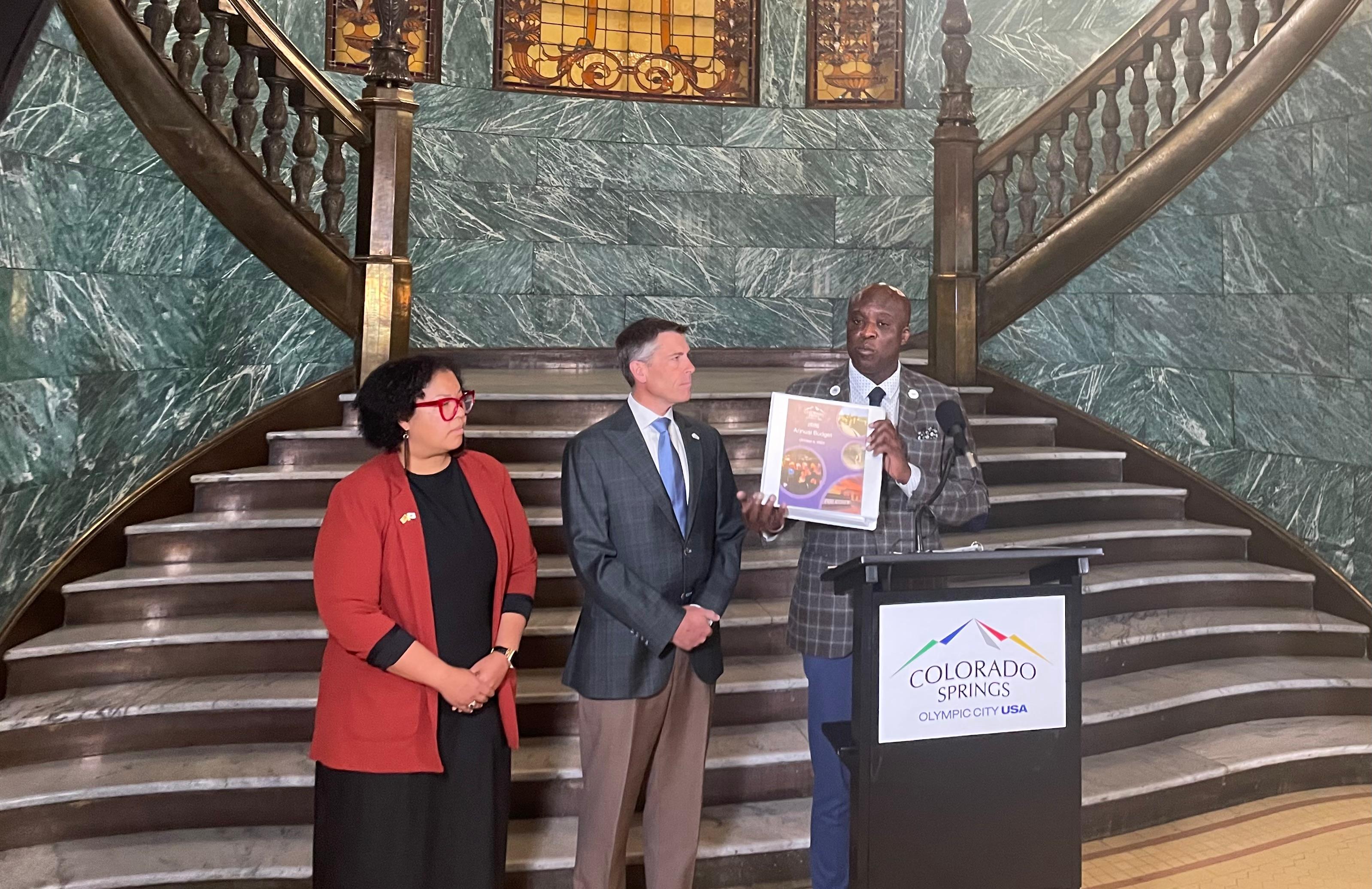 A man and a woman stand on the left with there attention to the man on the right who is standing at a podium and holding a large binder. Everyone is wearing buisness-wear. They are sitting at the bottom of two conjoining stair cases.