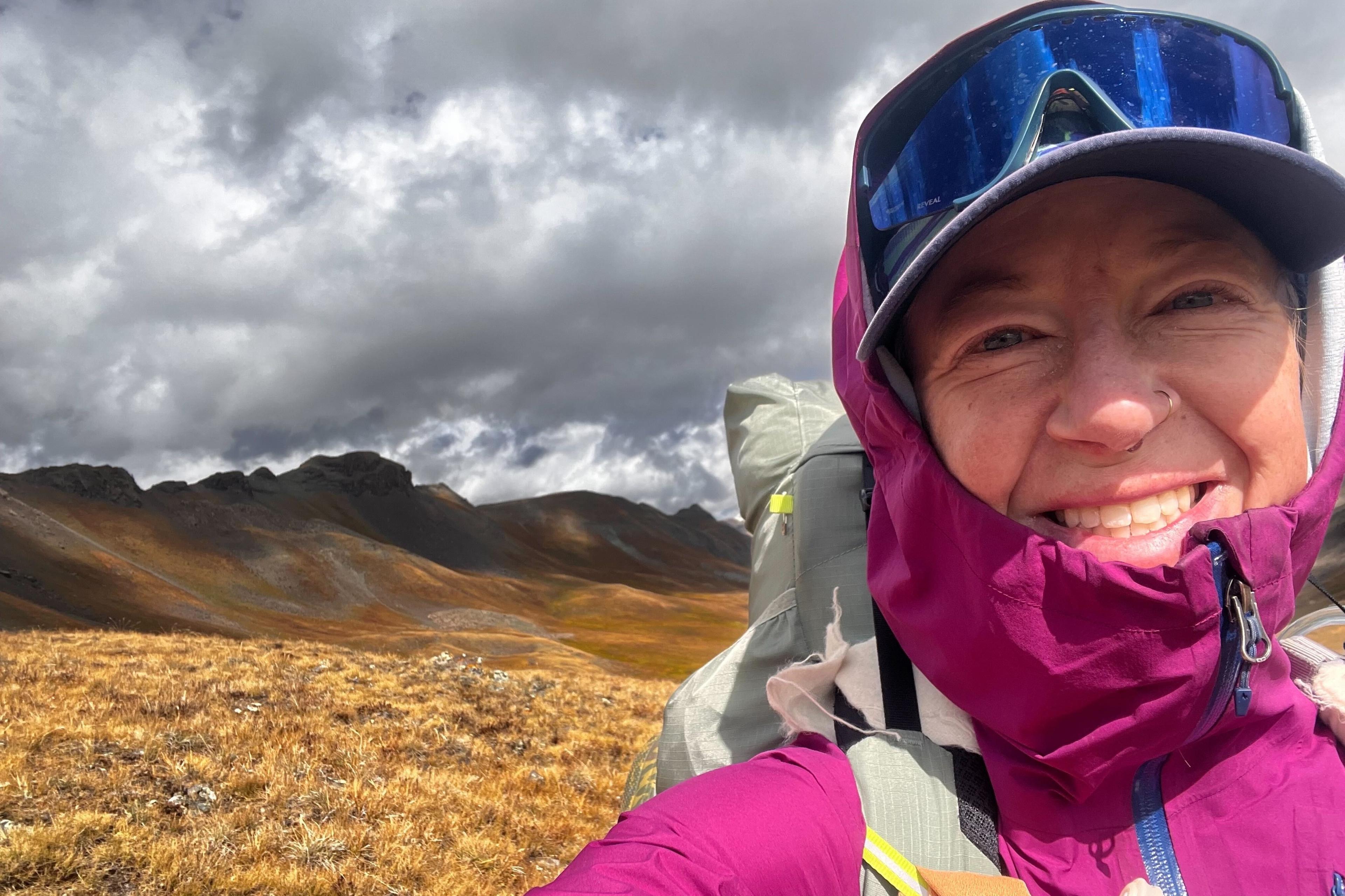 A woman in a pink jacket stands in a field with mountains and a cloudy background.