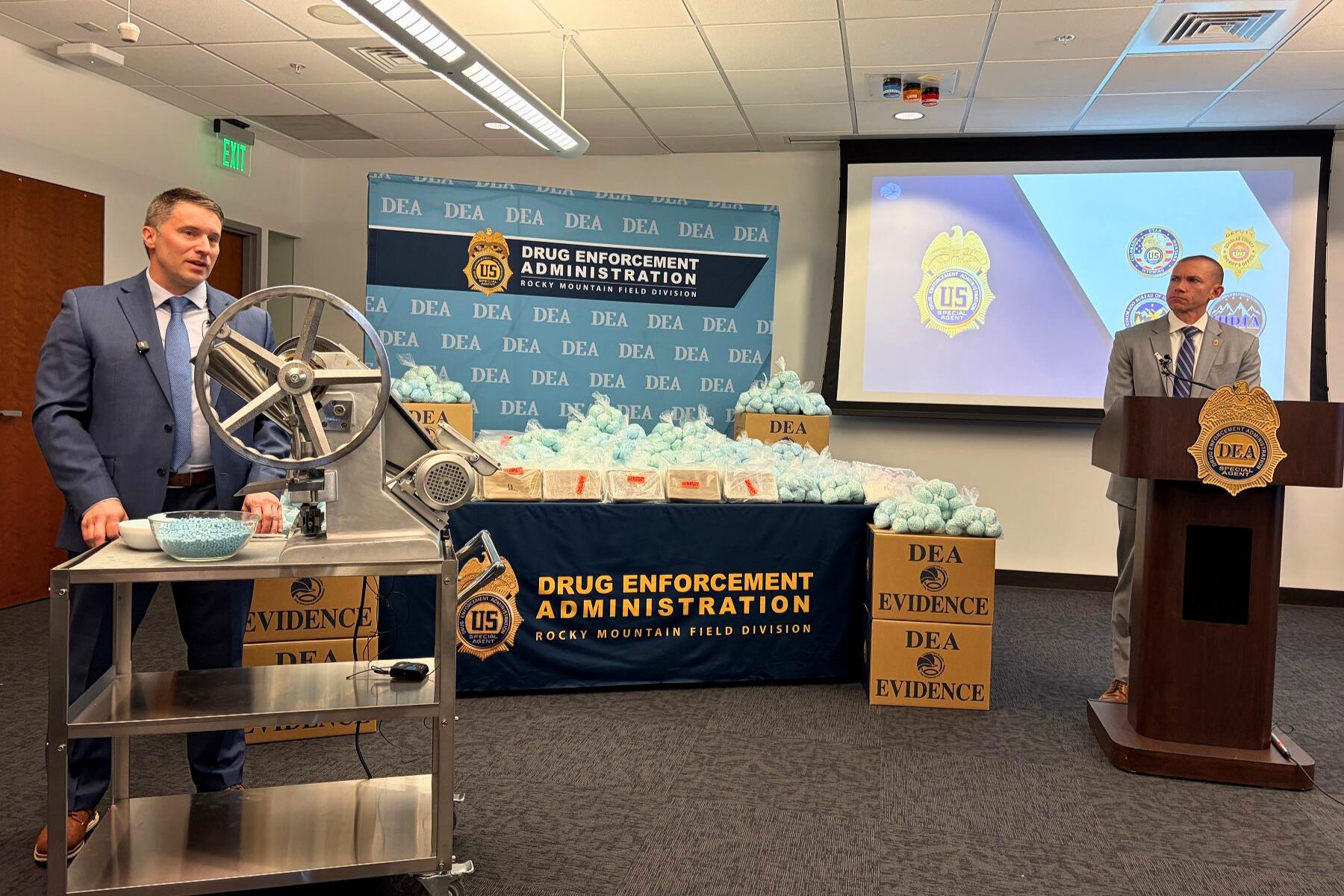 The image features a table with a blue tablecloth, topped with several boxes and bags. There is a person standing near the table, possibly attending to the items.
