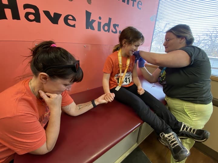 A nurse gives a young girl a vaccine in one arm as the girl, sitting on a medical bed, reaches across to hold her mother's hand with the other arm. The girl sitting on the table is wearing a number tag, possibly indicating a race or event.
