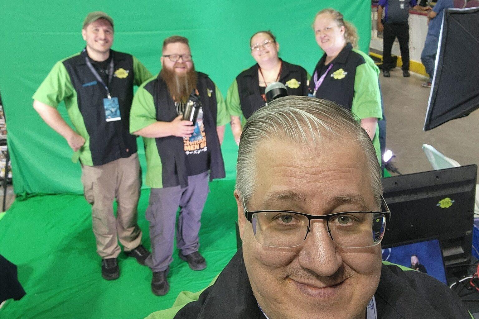selfie with man with gray hair and glasses in front standing in front of green screen with four people behind him.