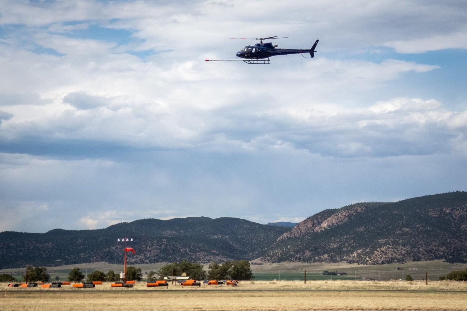 The image features a helicopter flying over a open area at an airport with mountains in the background. The helicopter is positioned towards the top center of the scene