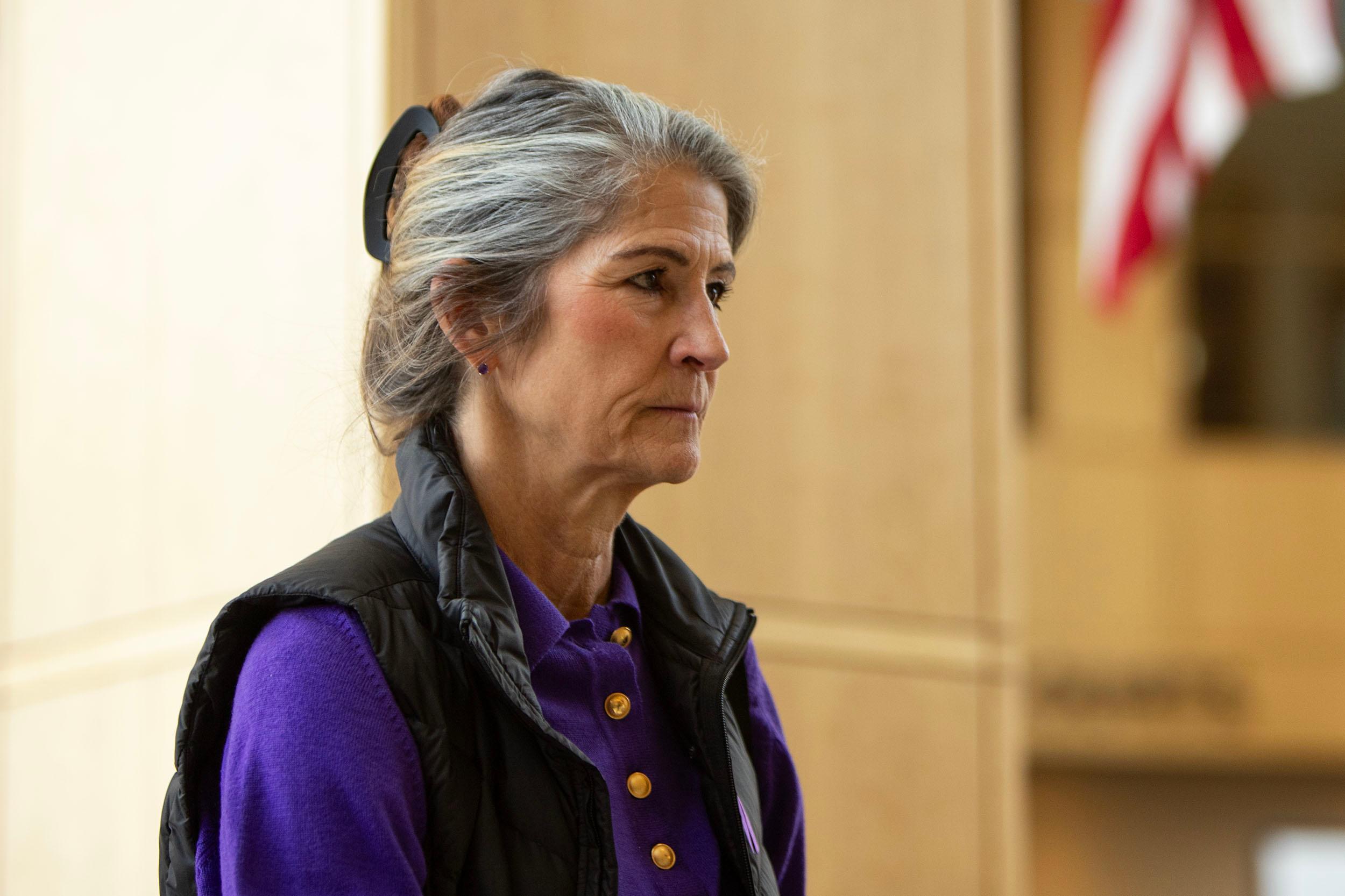 A woman in a purple shirt and a black vest stands in a warmly lit room.