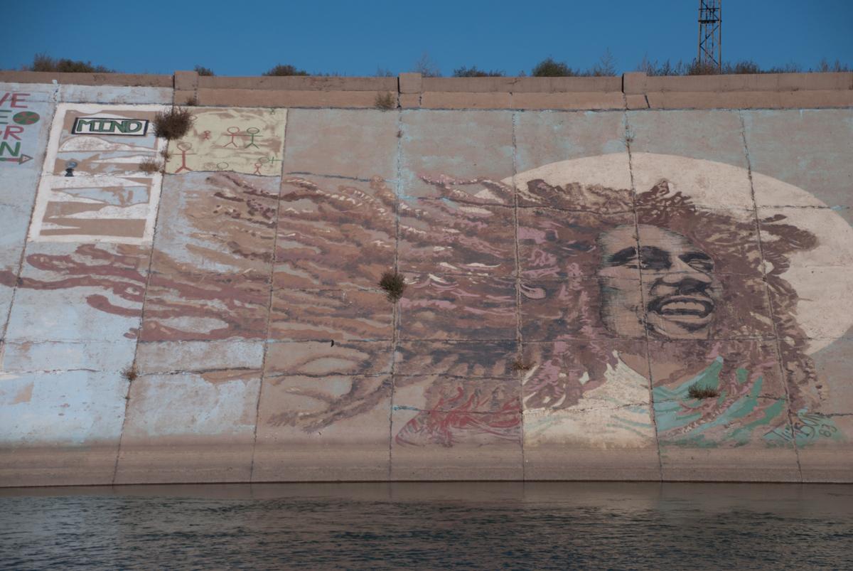 Photo of the same Bob Marley mural, taken Fall 2014, shows fading paint and plant growth through the concrete that makes up the levee.