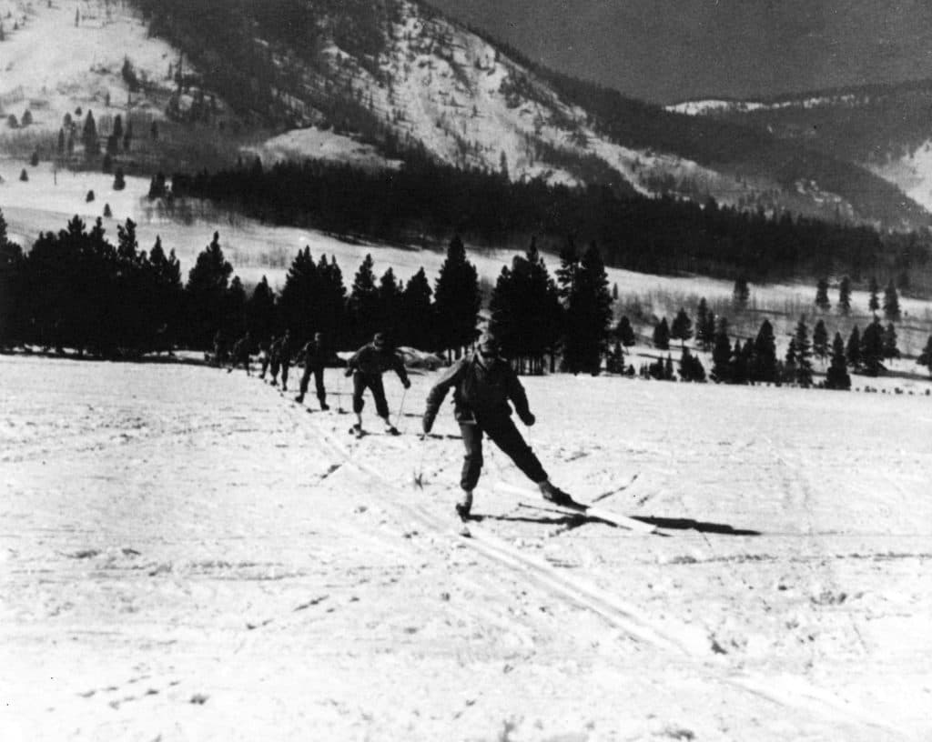 An undated historic black and white photo shows a line of soldiers on skis skiing toward the camera with mountains and pine trees in the background.