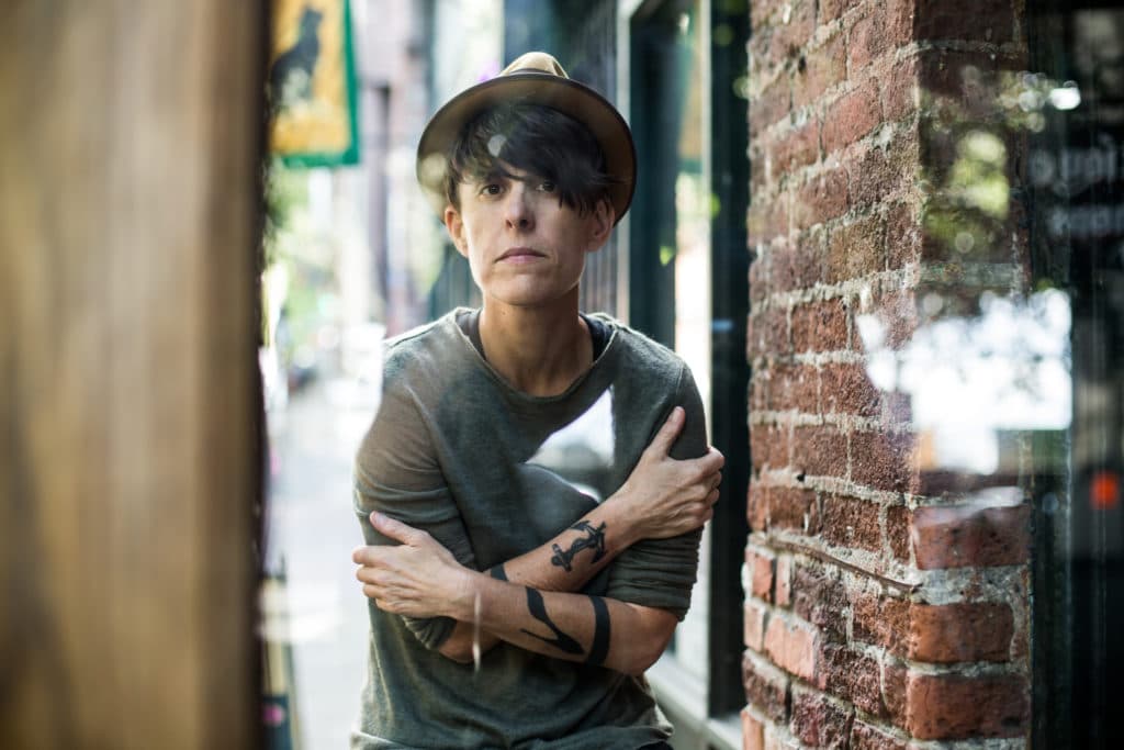 A person in a green shirt arms wrapped around their chest in a hat facing the camera brick wall to their left tree to their right