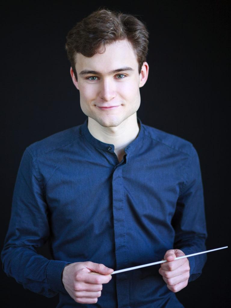 Assistant Conductor Bertie Baigent wearing a blue shirt, holding a batton, with a closed mouth smile.