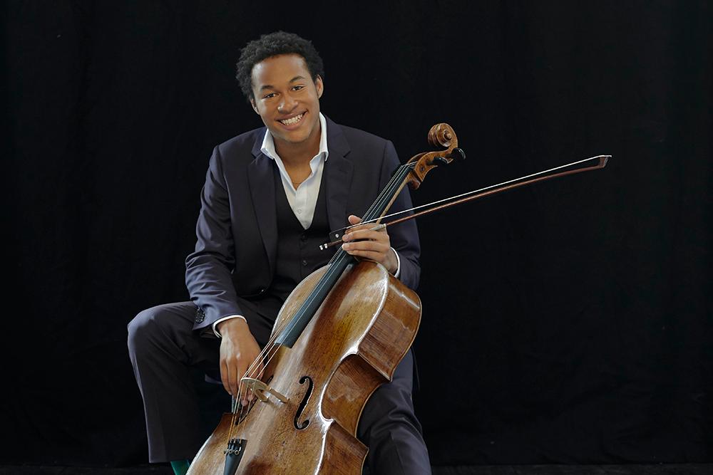 Sheku Kanneh-Mason sits holding his cello, smiling at the camera