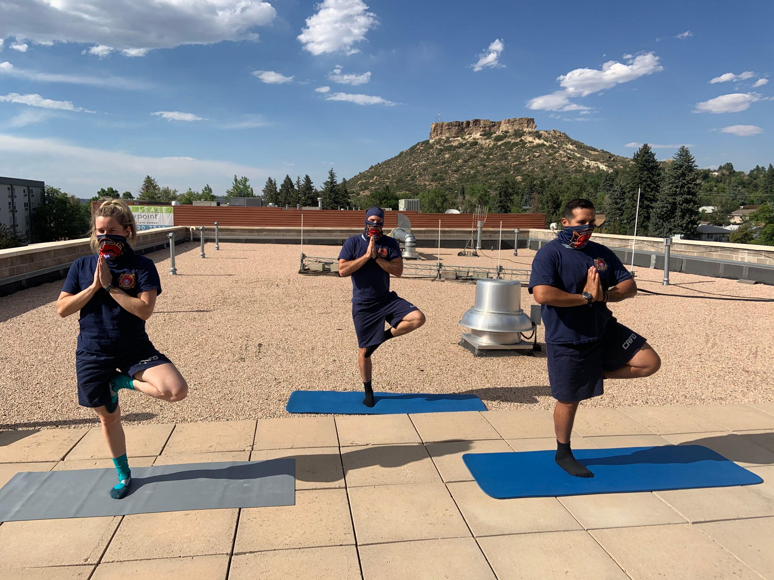 Three firefighters pose on a roof, doing yoga on blue yoga mats in masks.