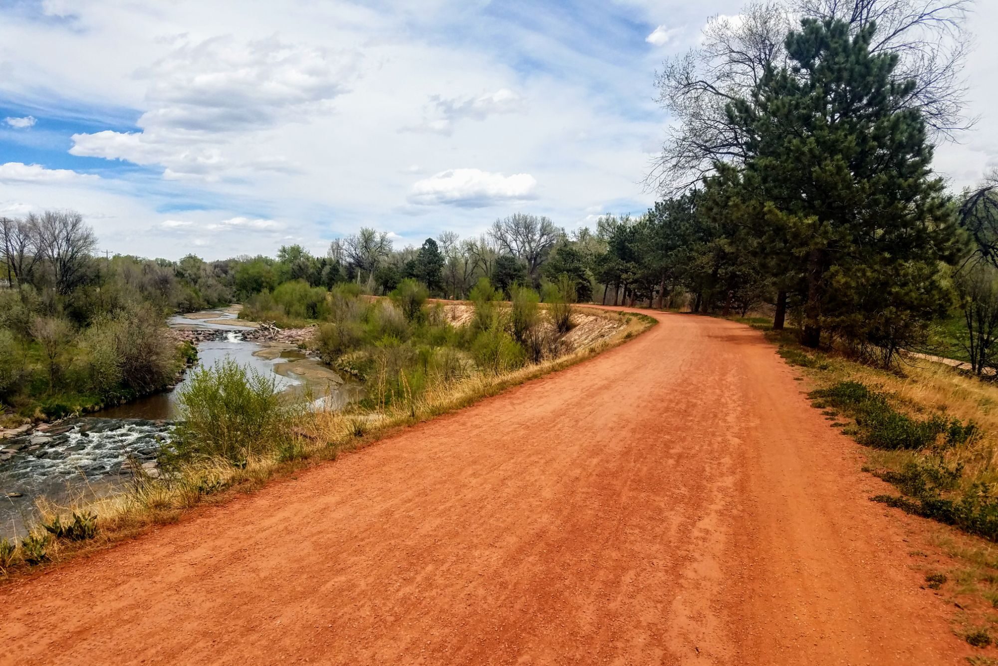 A long, red rusty bike path with trees in the distance and gray-green bushes to the left side.