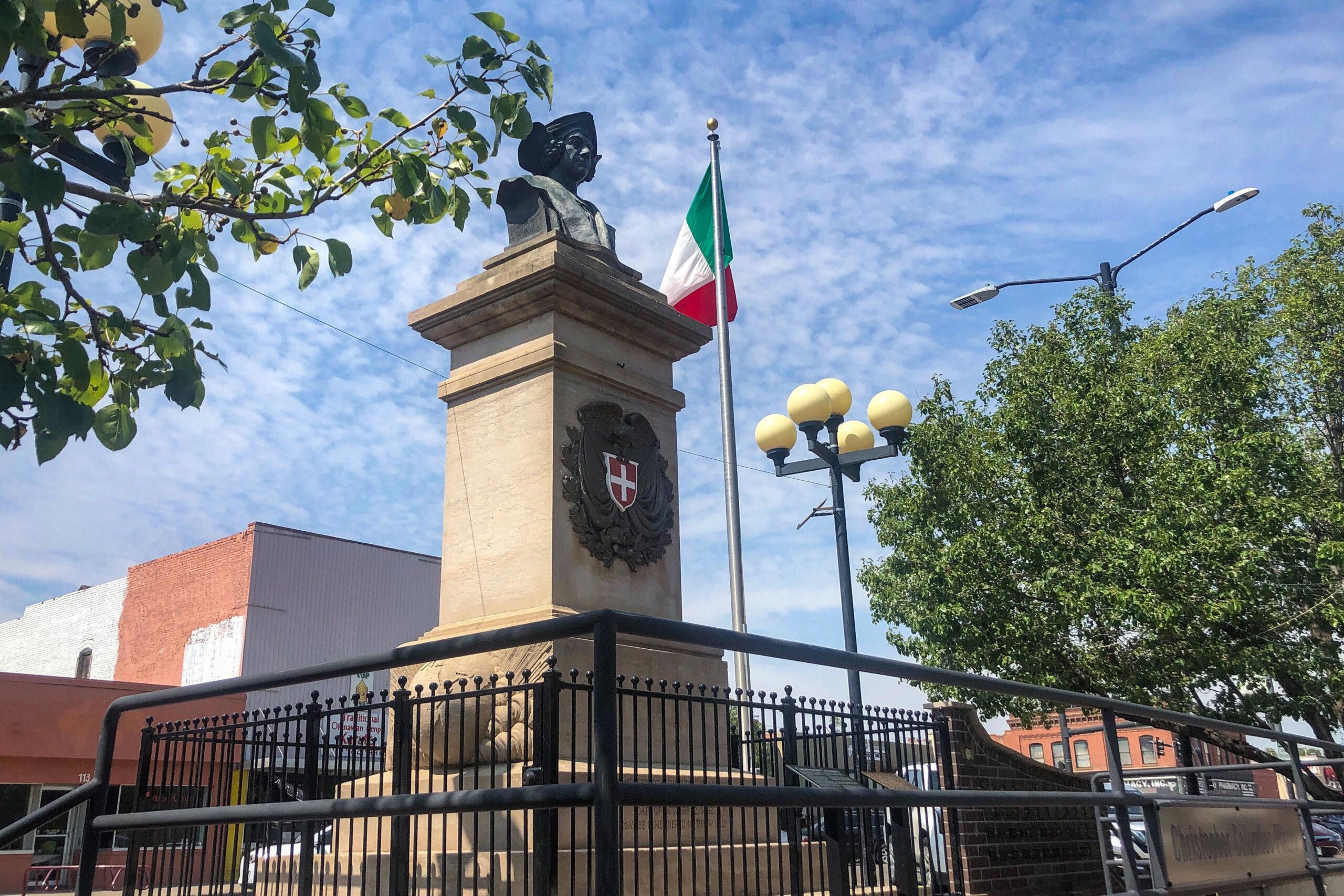 A view of the Christopher Columbus memorial in downtown Denver. A bronze bust of the explorer sits on top of a high stone pedestal, with an Italian flag in the background.