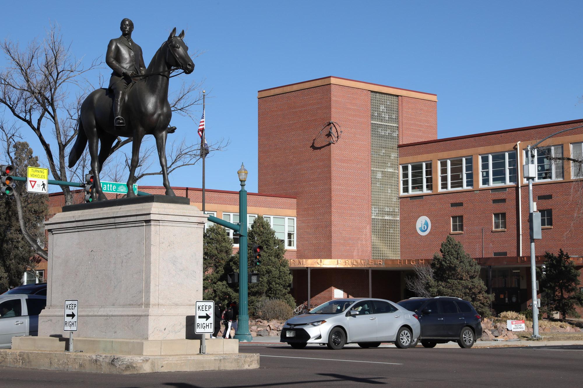 A bronze statue of a person on horseback stands on a stone pedestal at a street corner, with a red brick building and several cars at an intersection in the background.