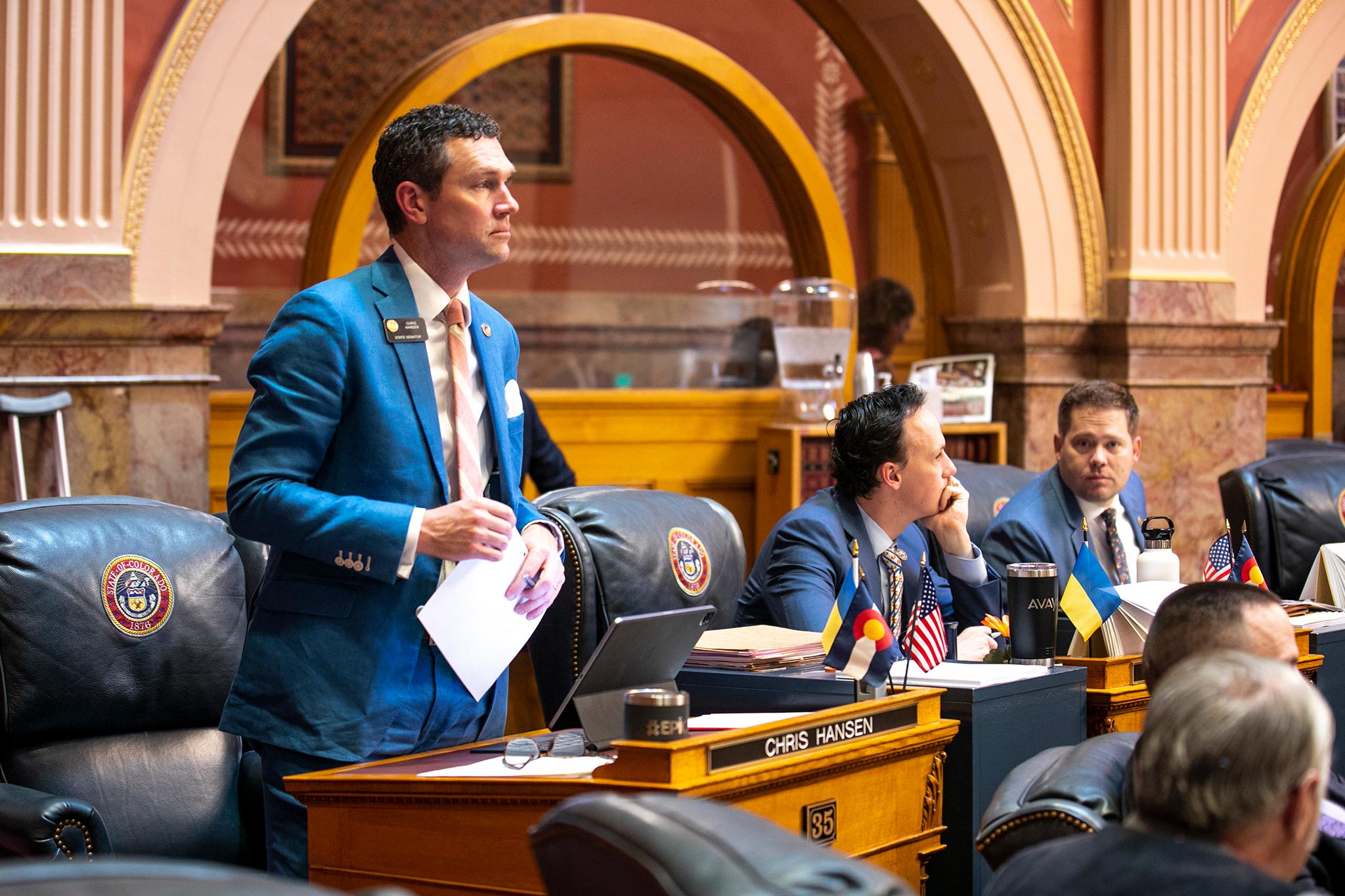 A man in a blue suit stands behind a desk in the Colorado Senate chamber, with two men seated at other desks beside him.