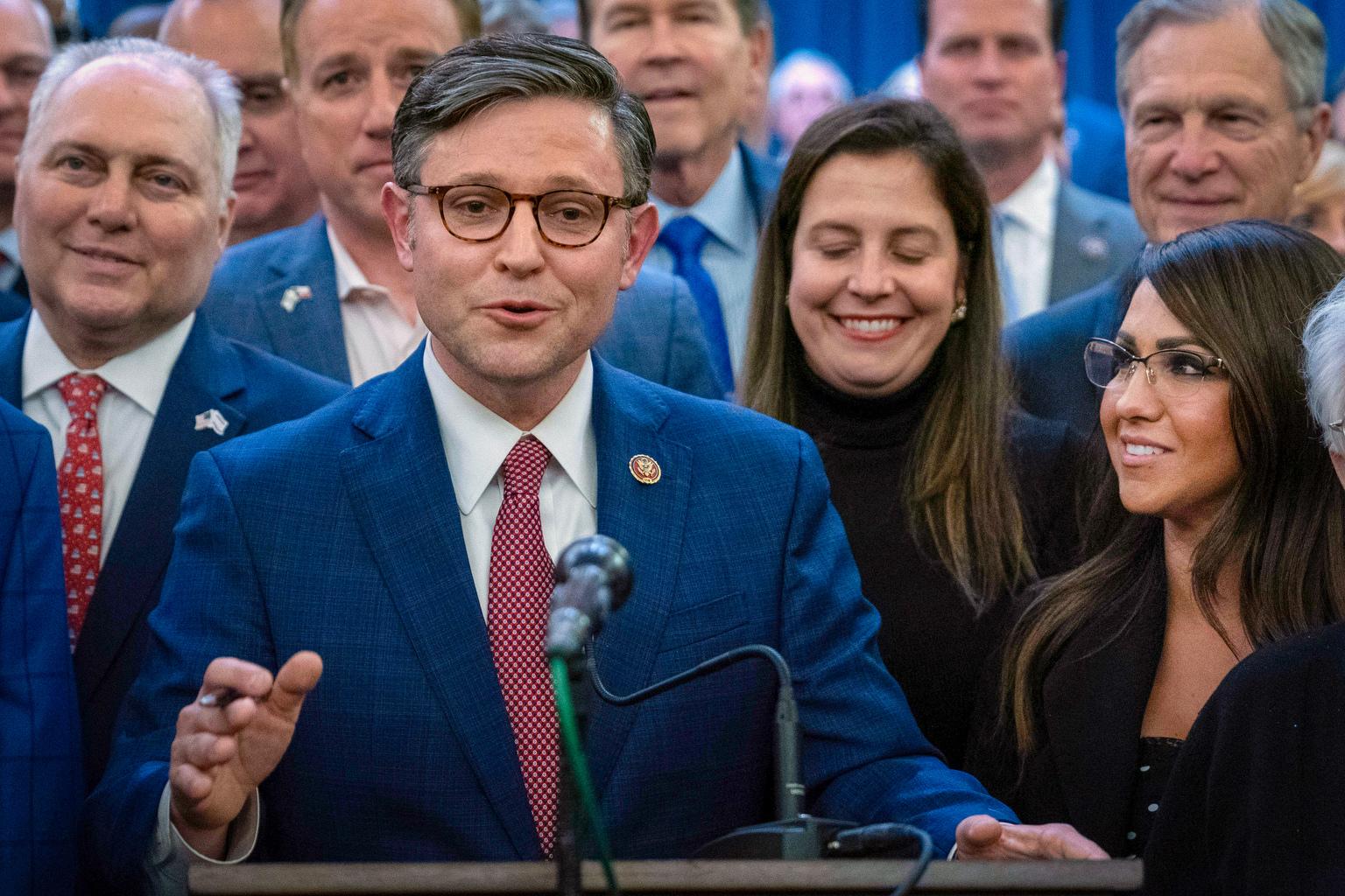 House Speaker Mike Johnson at a lectern with a crowd of people behind him and Rep. Lauren Boebert smiling at him from the side.