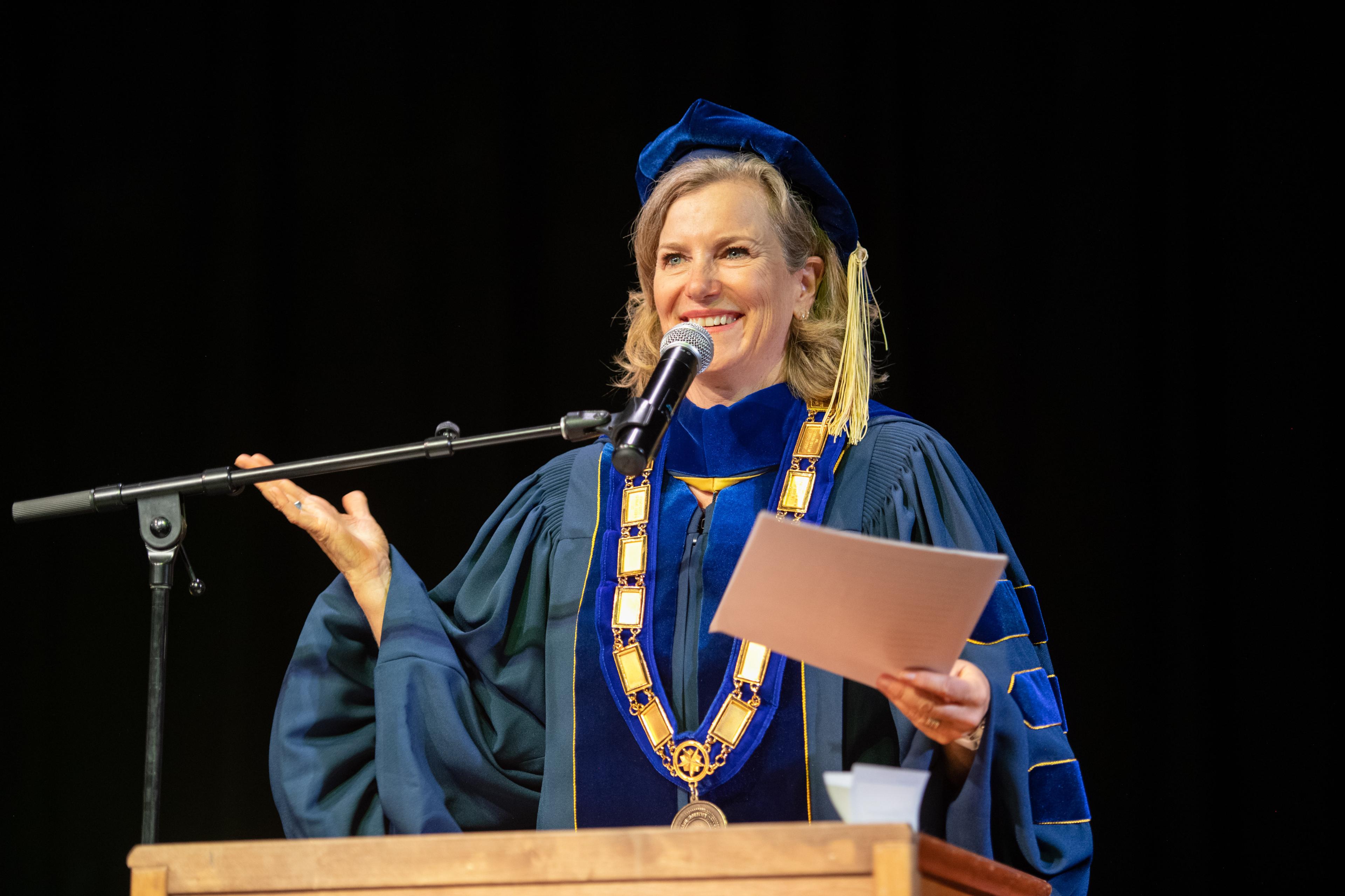 A female college president in blue robes smiles as she stands in front of a microphone.