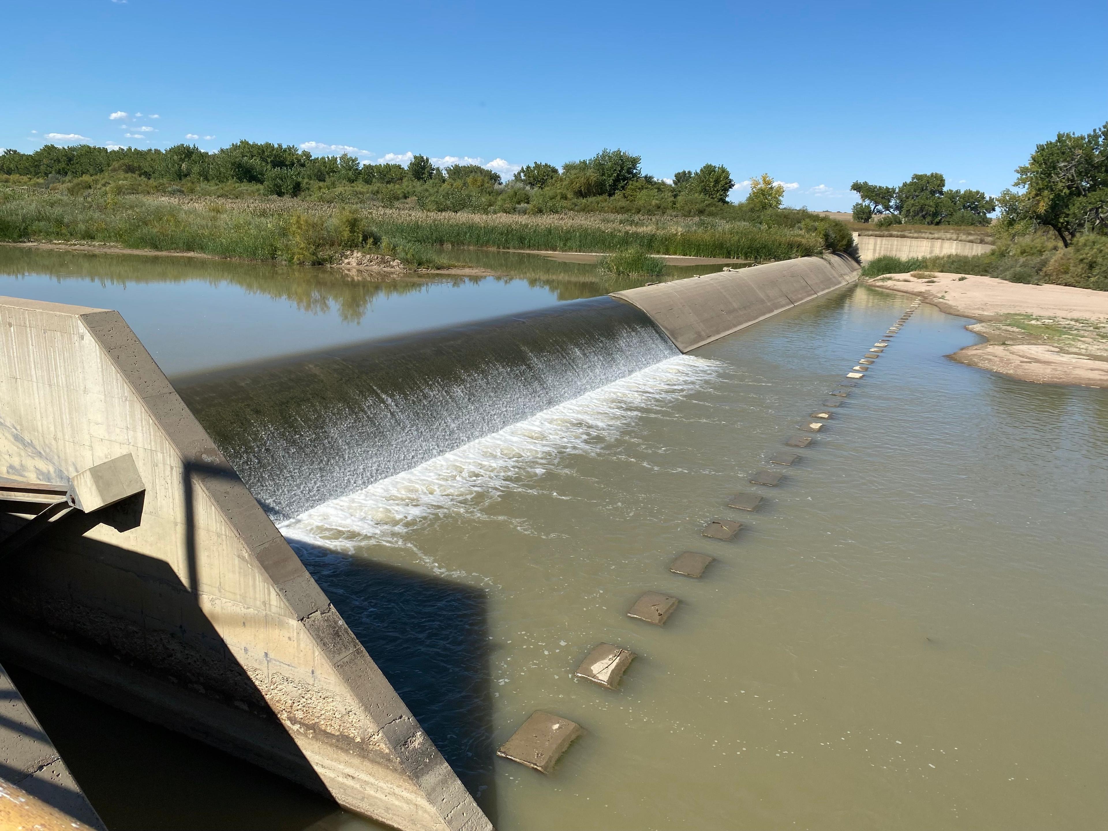 irrigation water flows over a small concrete dam with trees on the horizon and a cloudless blue sky above