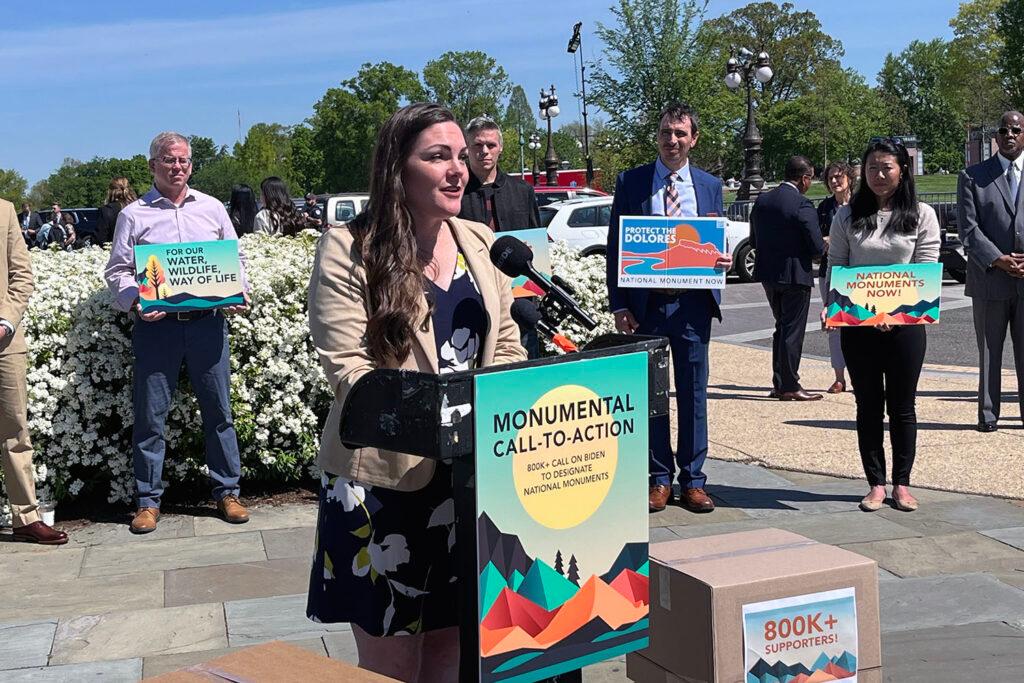 A photo of a woman speaking at a lectern that has a poster on it reading, &quot;monumental call-to-action&quot;