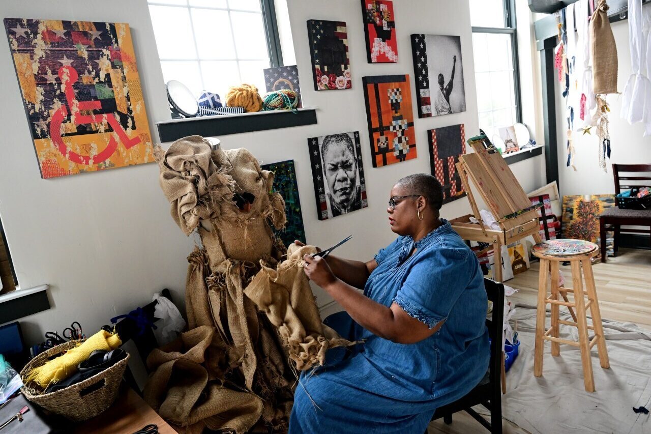 Denver artist Chloe Duplessis works on a fabric dress with a wall of bright paintings behind her.