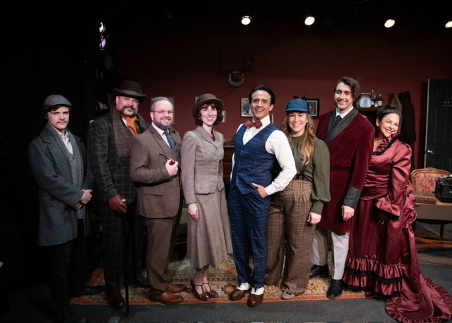 A group of actors in early 1900s attire pose on the stage where they perform.