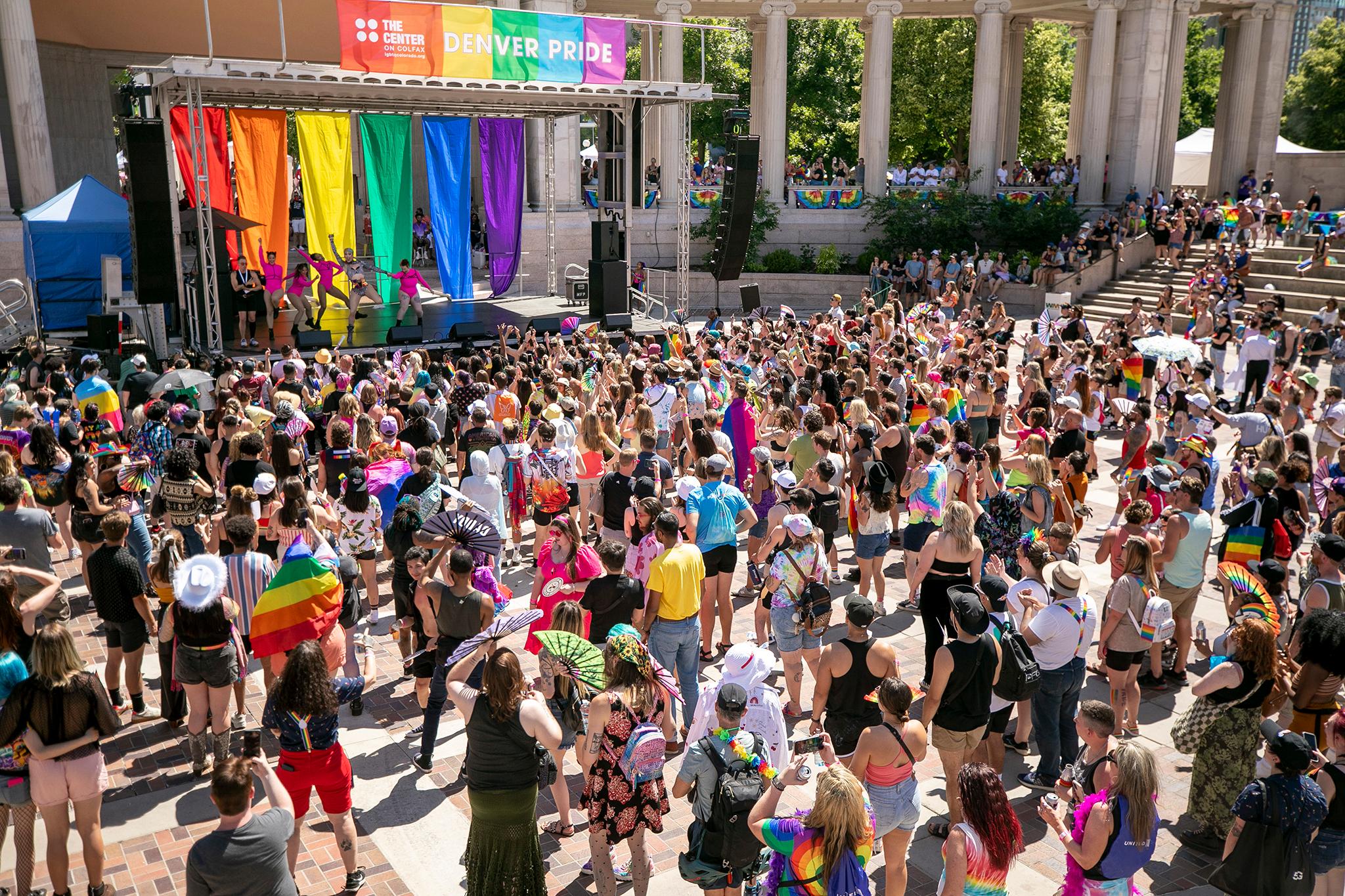 Crowds in Denver's Civic Center Park watch a musical performance on the main PrideFest stage with a background of rainbow banners.