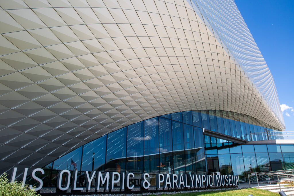 The exterior of the U.S. Olympic and Paralympic Museum. An arcing steel wall tops all-glass doors and windows on the first floor.