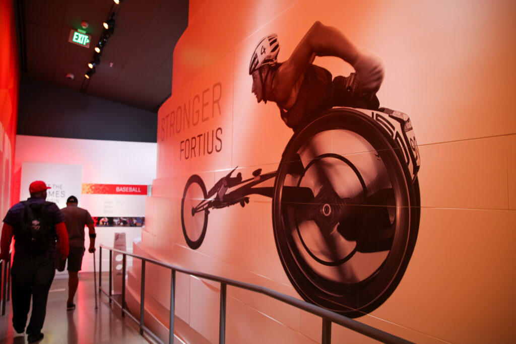 Two men walk down a ramp in the museum. The wall next has a mural of a Paralympic track athlete.