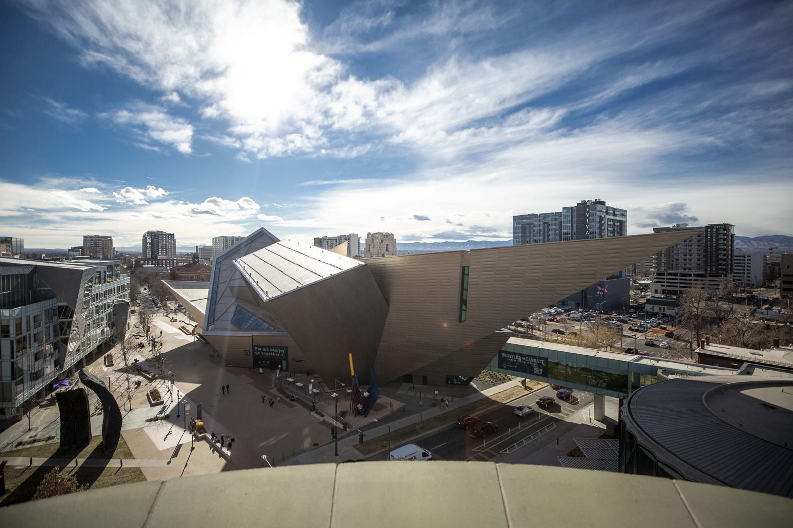 A view of the Denver Art Museum from Denver Public Library's Central branch. The DAM is a grey big building with angular sides that jut out in different directions. It's surrounded by other buildings, roads, and cars.