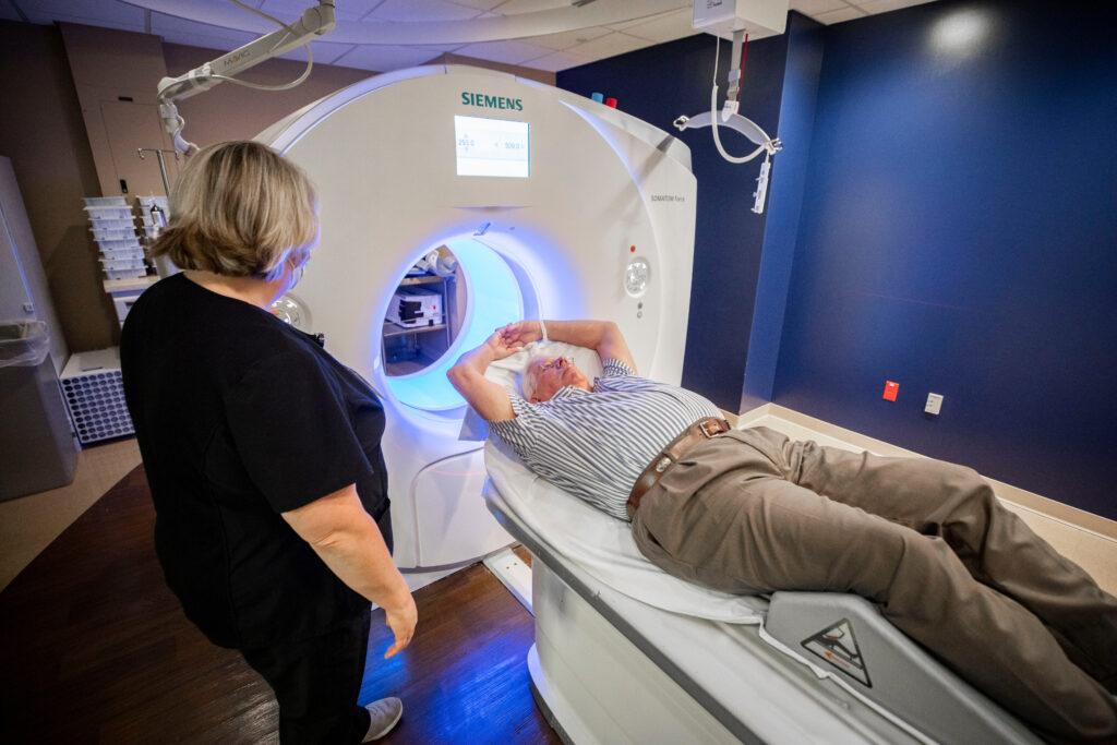 A man lays on a CT machine table with his arms over his head as a nurse operates the medical machine.