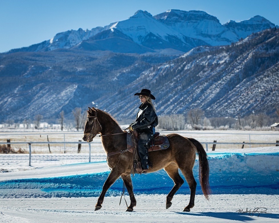A woman sits on a horse in the snow. The mountains are in the background.