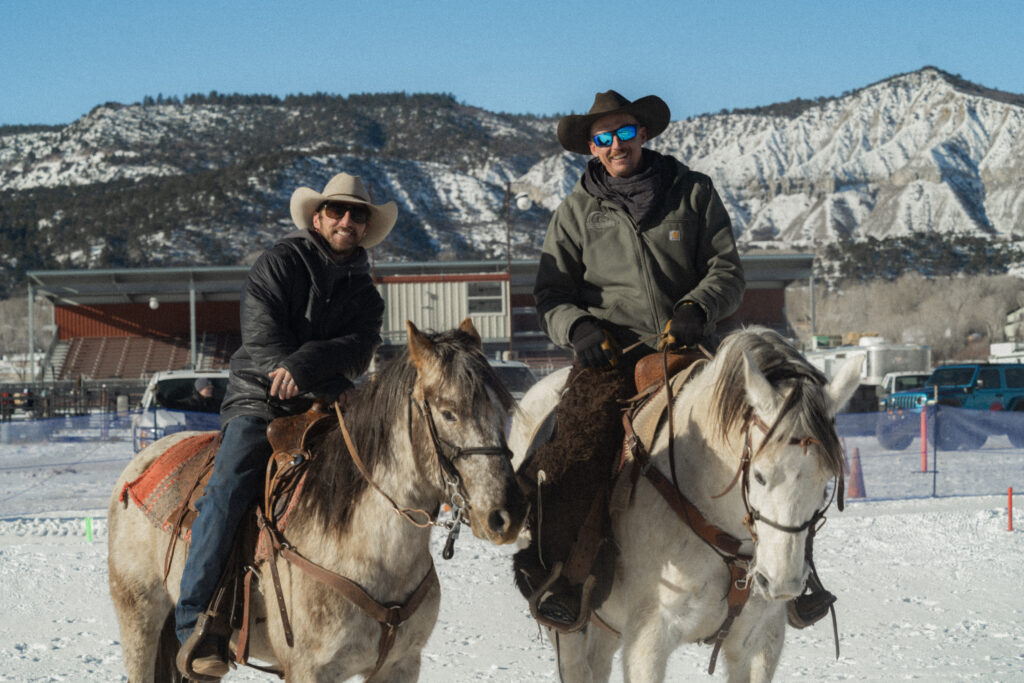 Two men sit atop horses in front of an arena grandstand where skijoring races take place in the snow.