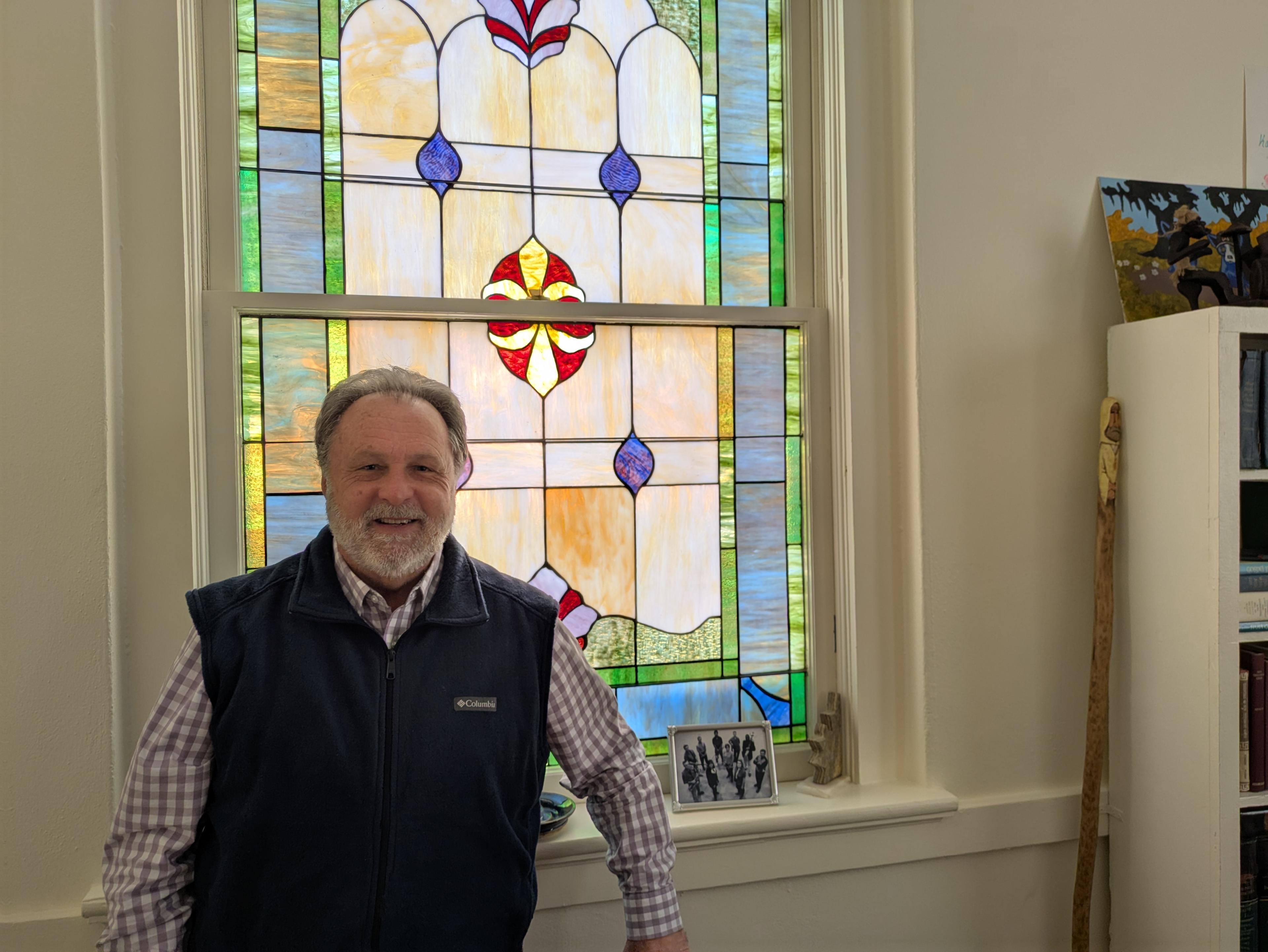 Rev. Kevin Young, wearing a checkered dress shirt and blue vest, stands in front of a stained glass window.