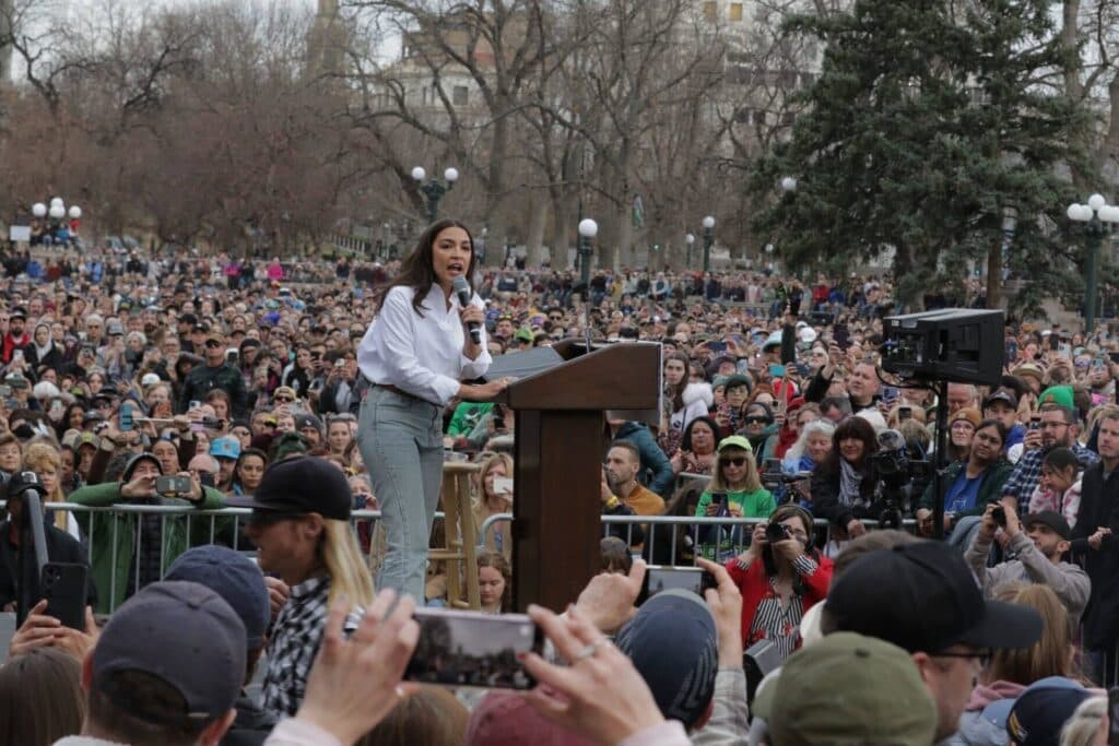 Alexandria Ocasio-Cortez speaks to a large crowd as people take photos on their phones.