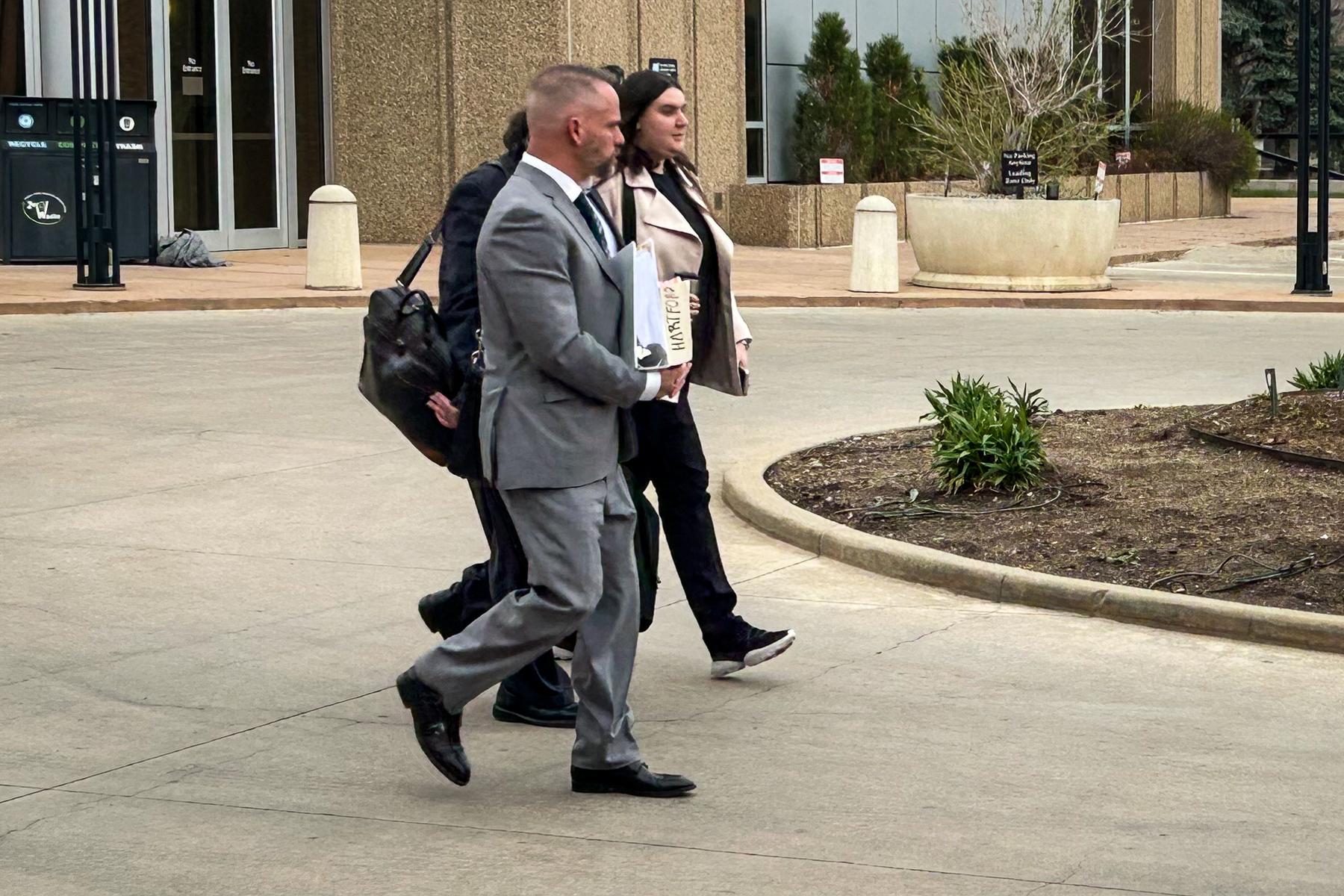 Three people are walking in profile to the right. The one in the foreground is a man in a gray suit.