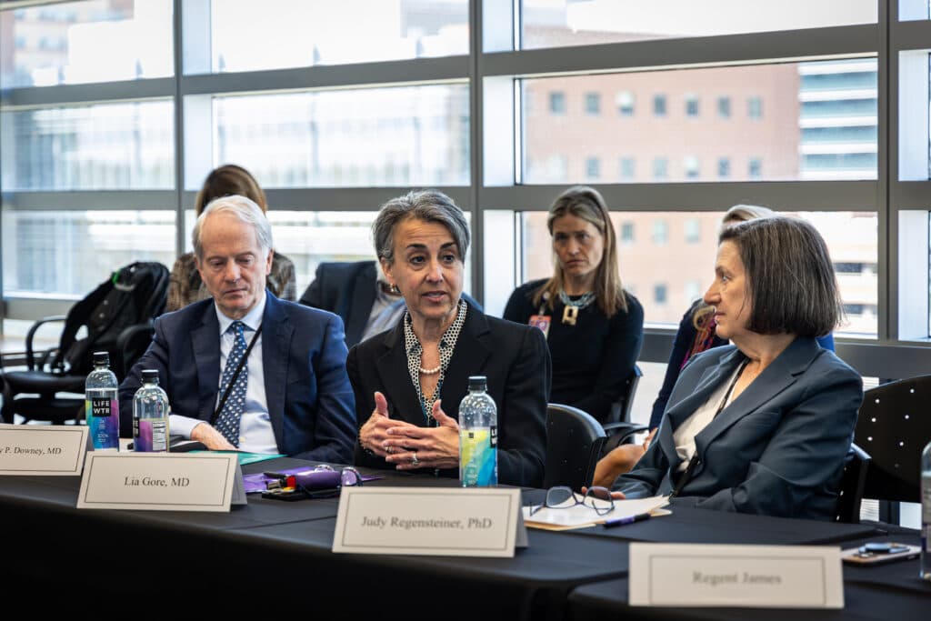 In the foreground, a man and two women sit behind their name plates at a conference room table. The woman in the middle is speaking and holding her hands out in gesticulation. There are several people in the background listening.