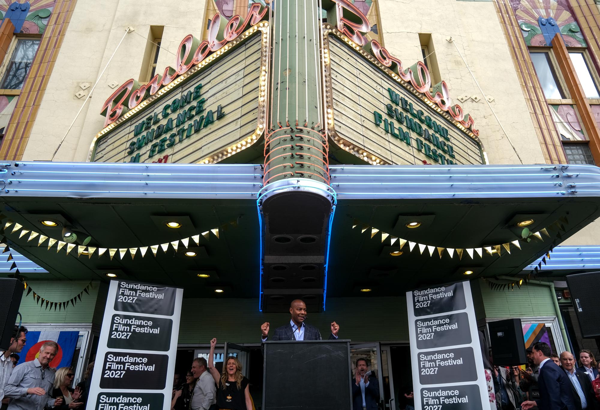 A man stands at a lecturn in front of a big theater marquee.