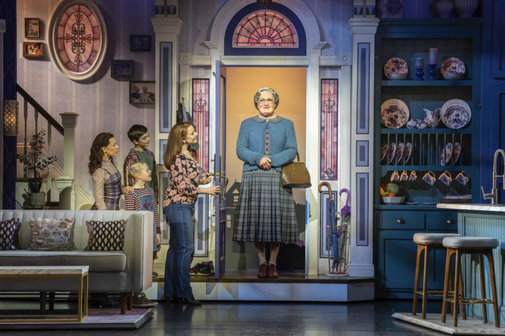 A still from the Broadway production of Mrs. Doubtfire, showing characters opening the door to their home with Mrs. Doubtfire on the stoop, hands crossed over her stomach and a slight smile on her face.