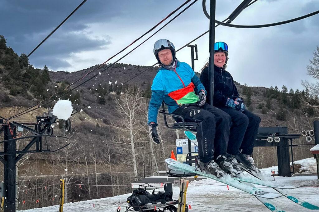 A man throws a snowball off a two-seat ski lift as a woman next to him smiles.