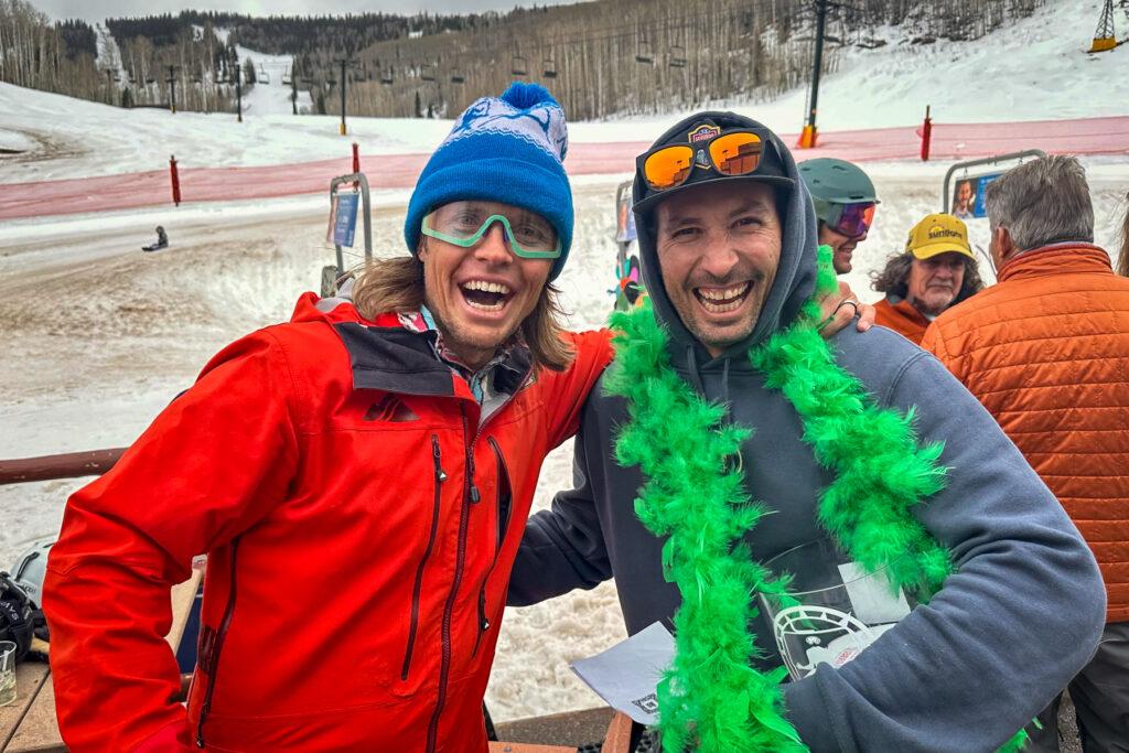 Two happy men embrace and smile with a historic ski lift behind them.