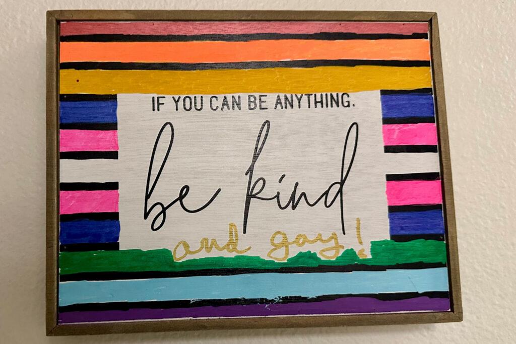 With rainbow colors behind the text, a painting says: If you can be anything be kind — and gay!