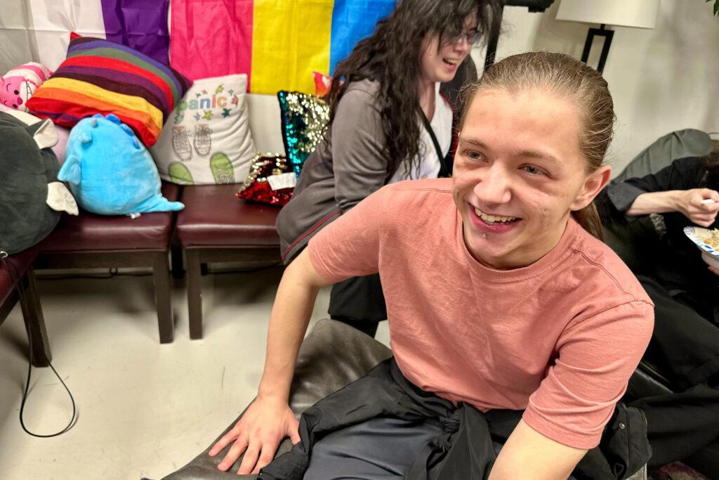 A young non-binary person with long hair smiles in front of people eating.