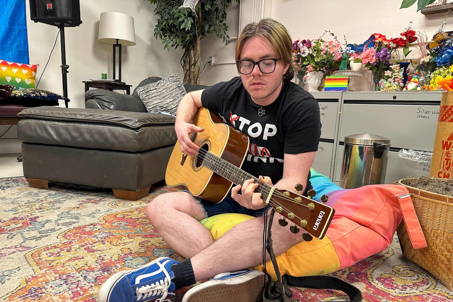 A young transgender man plays guitar as he sits on a rainbow pillow, with flowers and pride flags behind him.