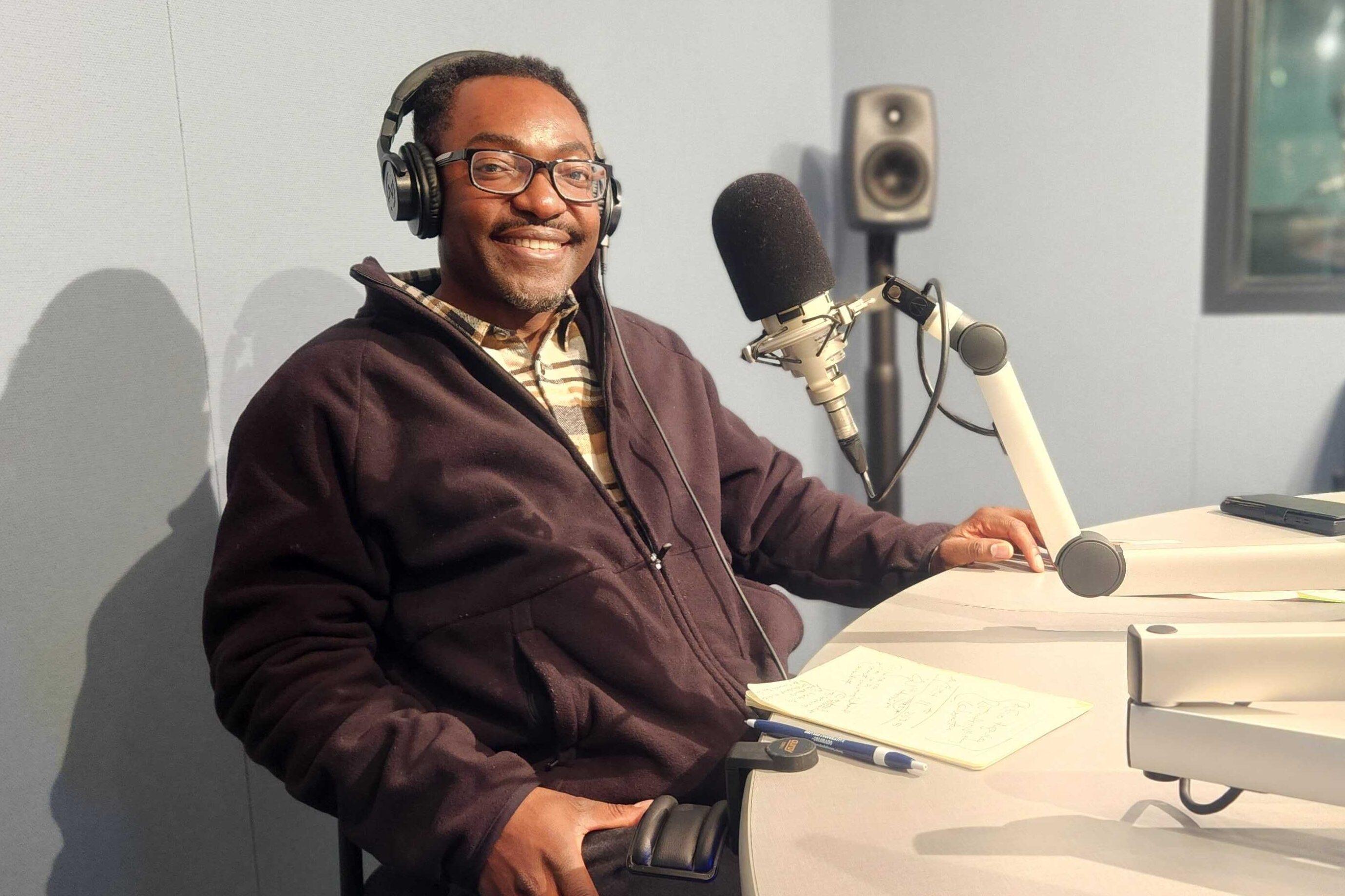 George Bamu is seated in front of a microphone. He is wearing headphones and is smiling at the camera.