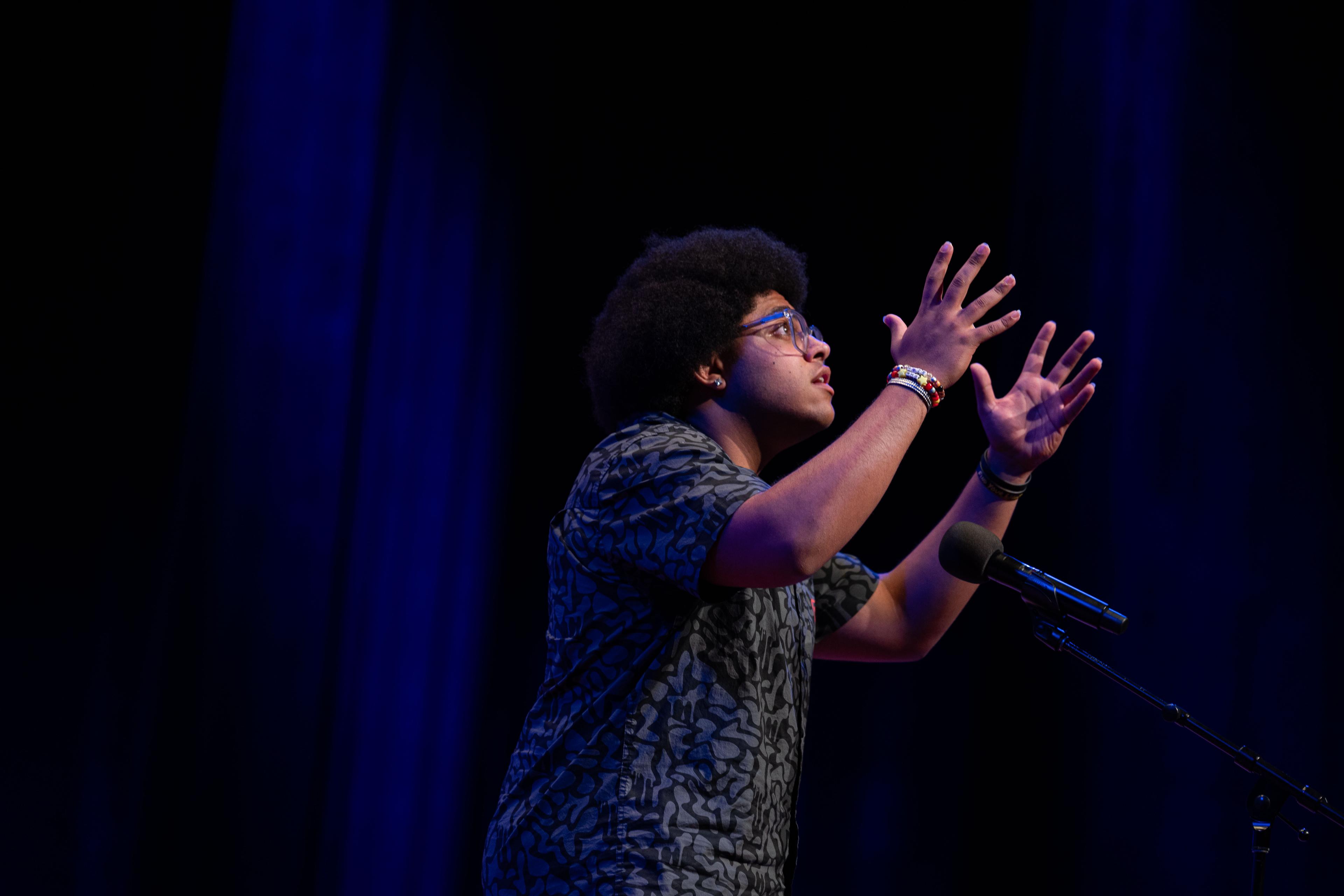 A student is pictured from the side with his hands raised in the air as he recites a poem on a dark stage.